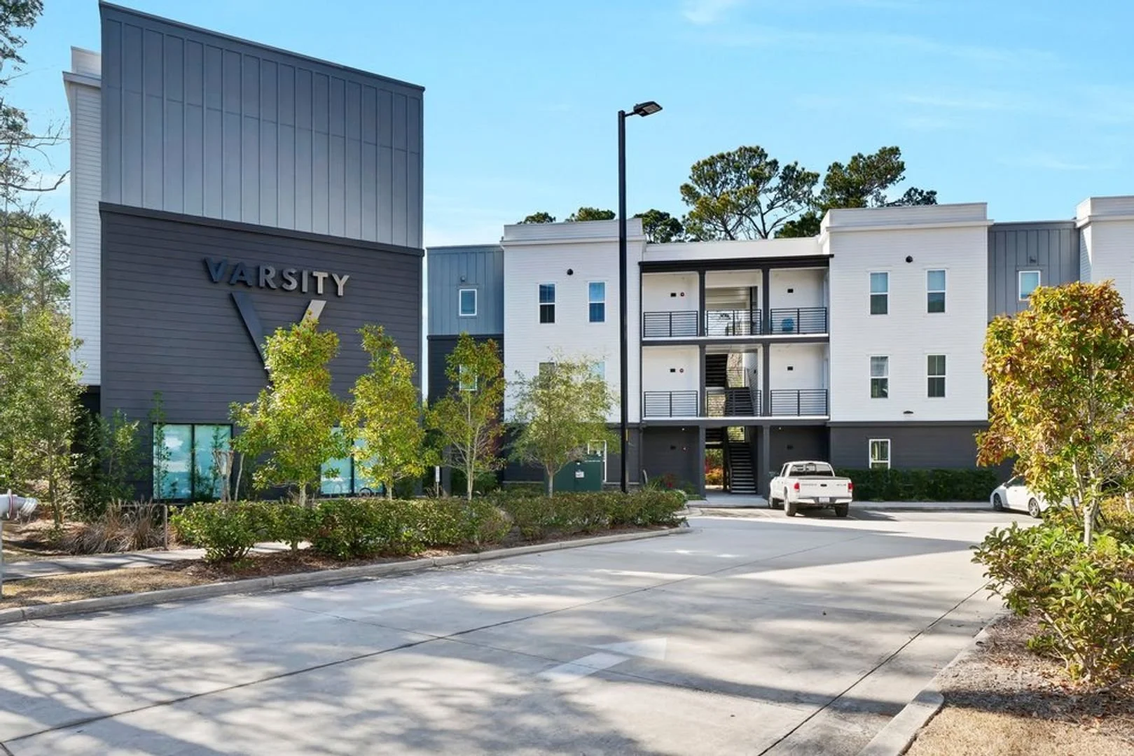 A modern apartment complex with a sign that says 'VARSITY'. The building has white and dark gray sections with multiple balconies. There are several trees, bushes, and parked cars in the parking lot, with a clear blue sky overhead.