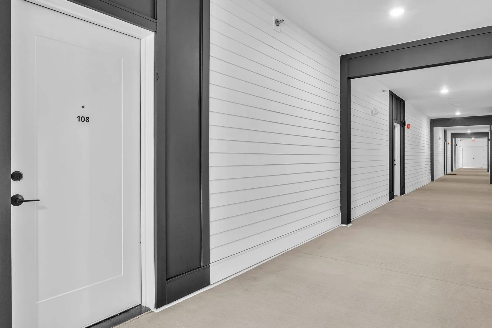 An indoor apartment corridor with a white door marked 108 on the left and multiple doors along the hallway, with beige flooring and white paneled walls.