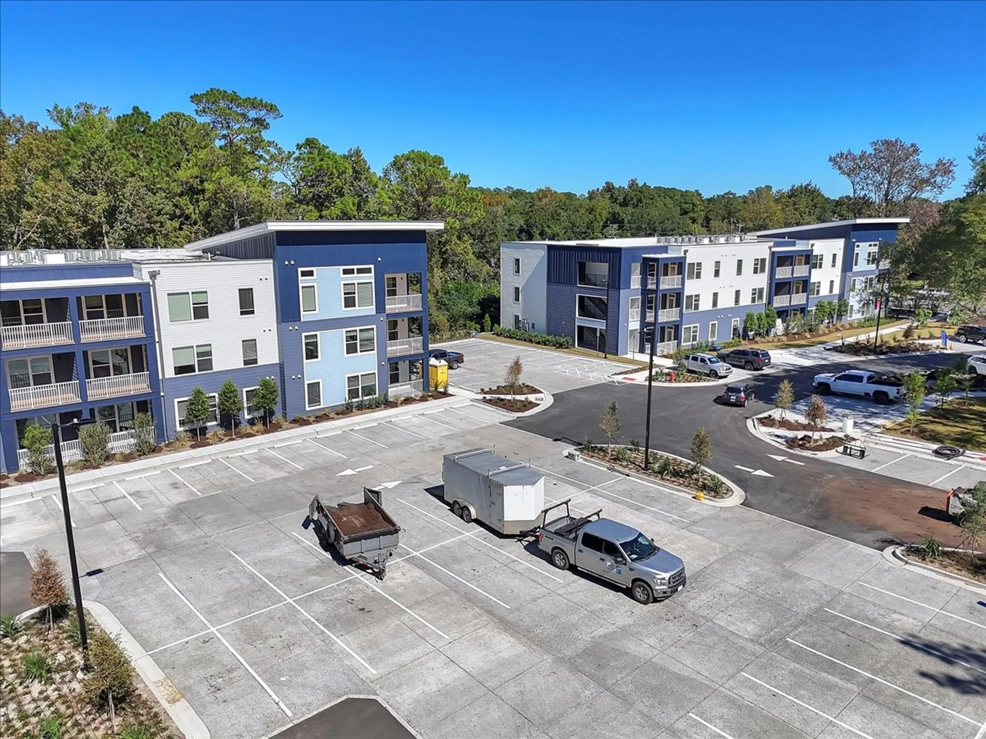 A large parking lot with few vehicles, surrounded by modern apartment buildings painted in shades of blue and white, with some trees and landscaped areas, under a clear blue sky.