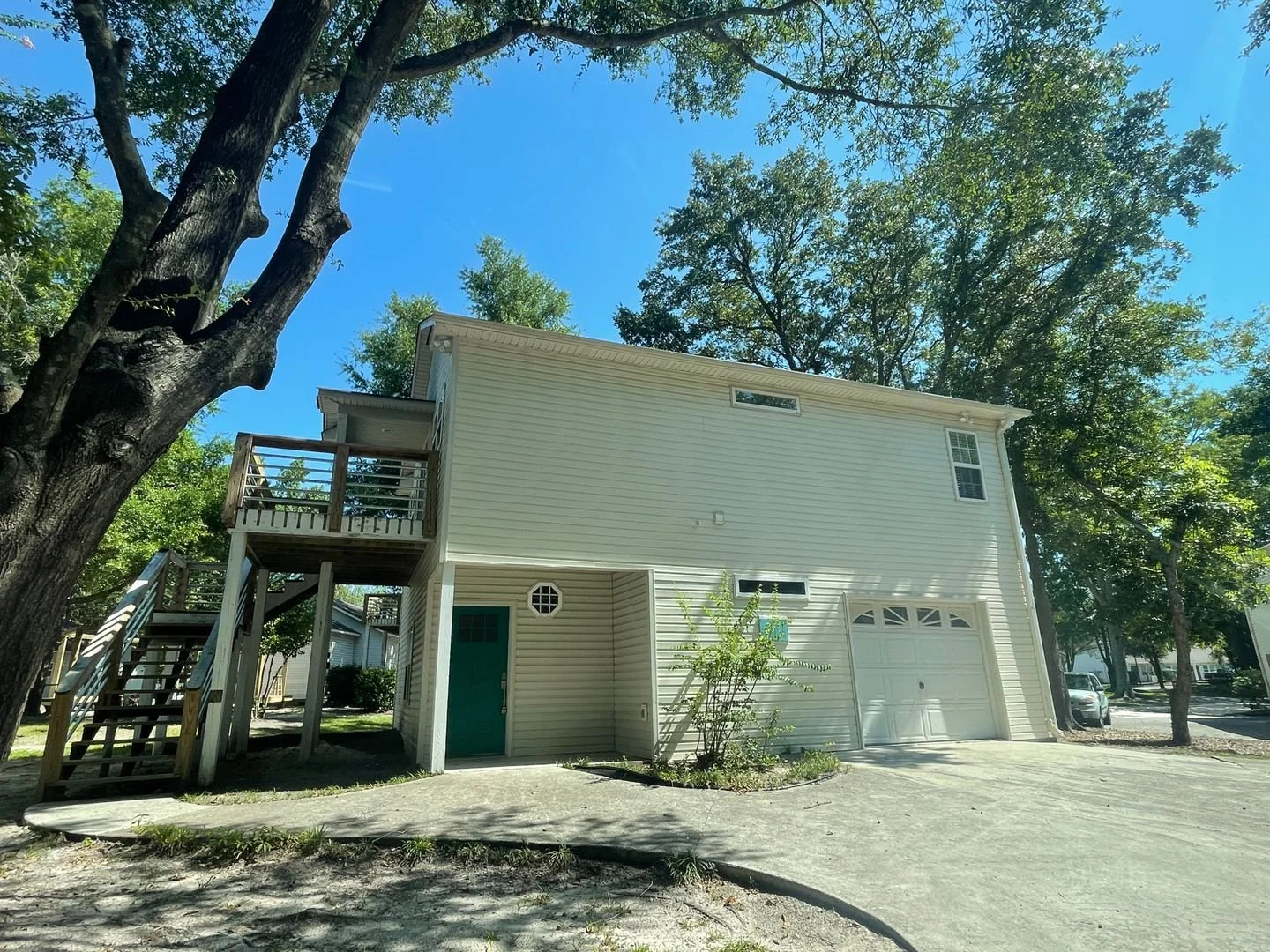 A beige multi-story house with a green door, a small front yard with bushes, and a driveway, surrounded by tall trees under a clear blue sky.