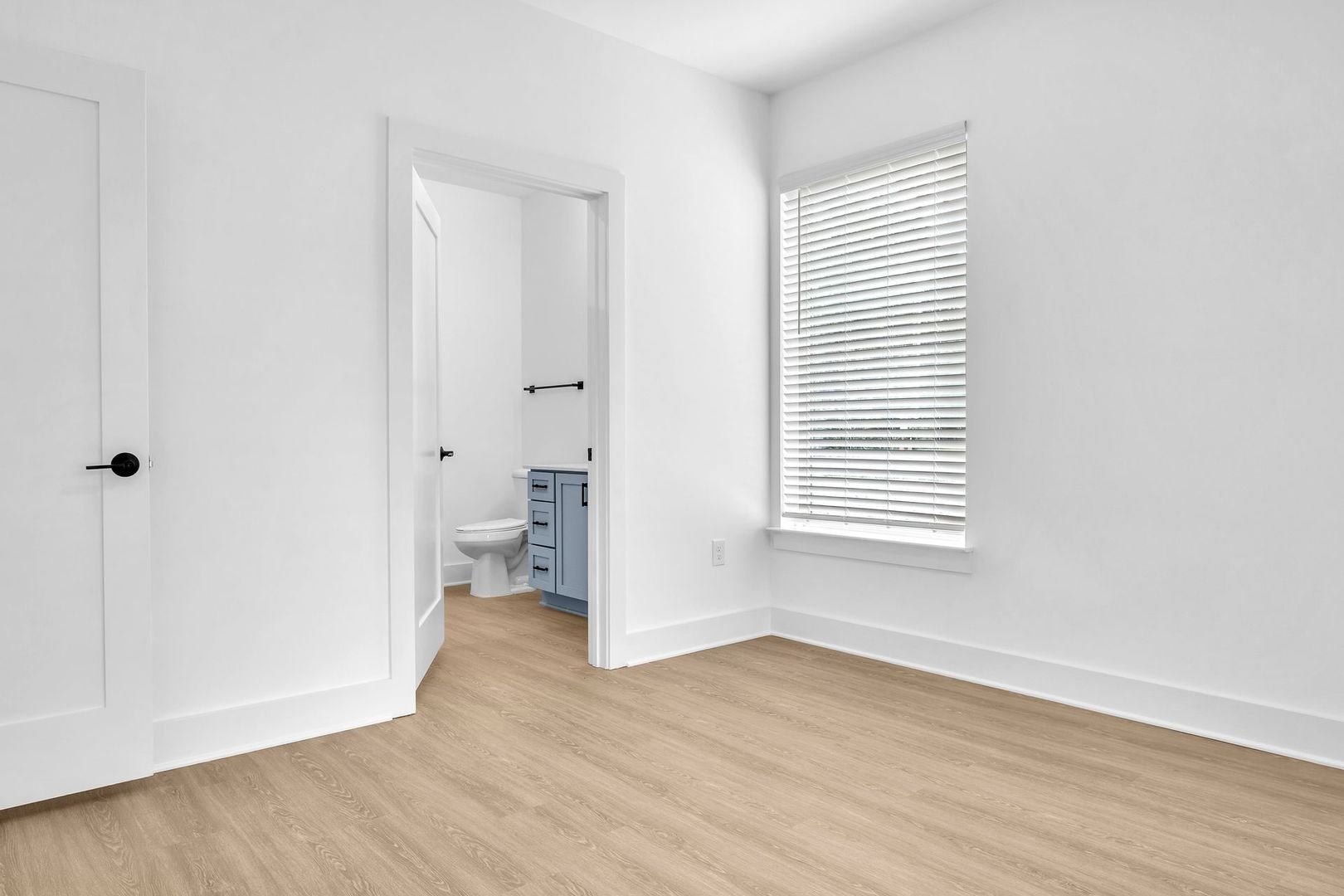 Empty room with beige wood floor, white walls, window with closed blinds, and an open door leading to a small bathroom with a toilet and blue vanity.
