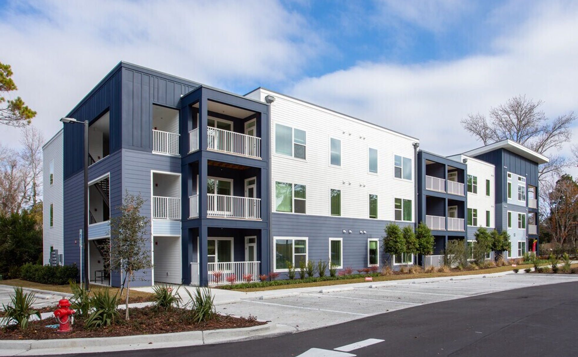 Modern multi-story apartment building with balconies, white and blue exterior, landscaped surrounding, and parking lot in front.