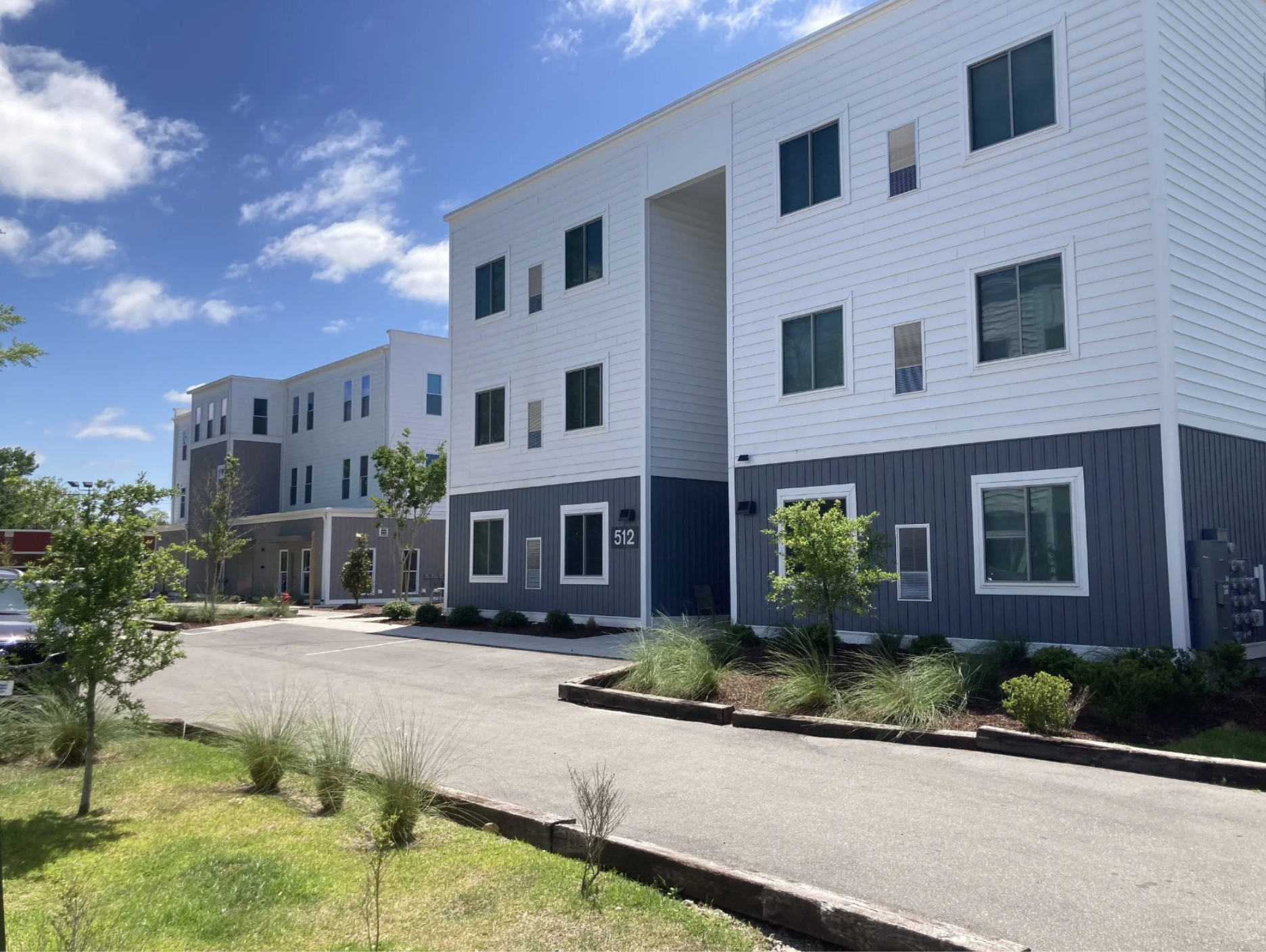 New modern apartment building with white and dark gray siding, multiple windows, and a leafless tree in the foreground under a clear blue sky.