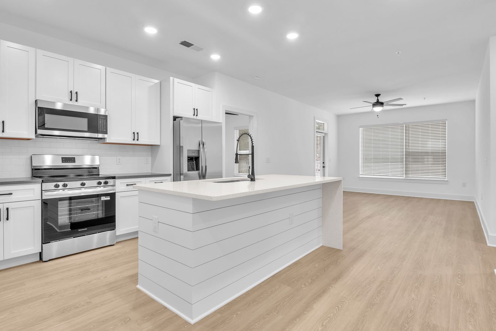 Empty kitchen with white cabinets, stainless steel appliances, a large island with a sink, light wood flooring, and a living area with windows and a ceiling fan.