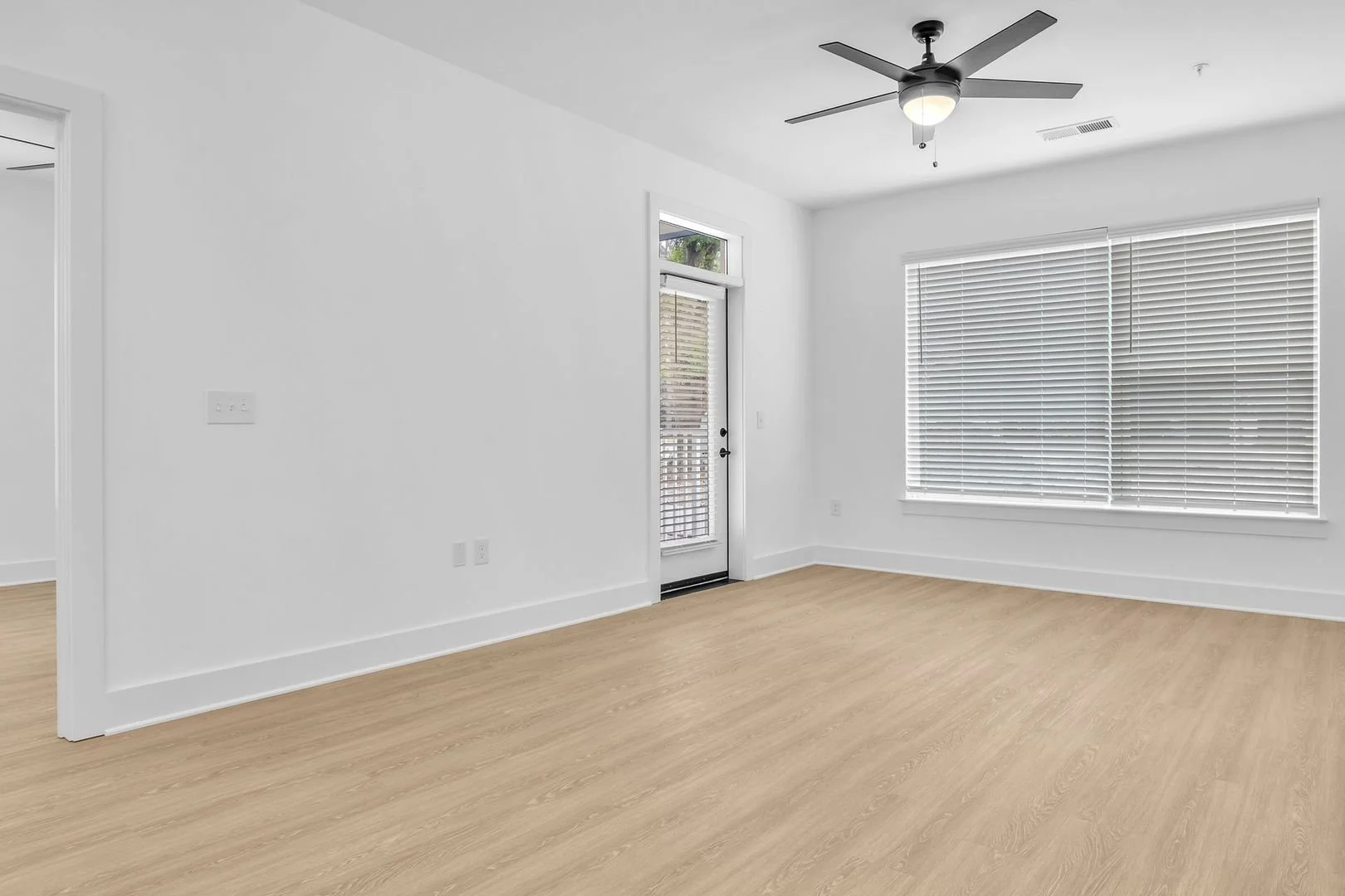 Empty bright living room with white walls, light wooden floor, large window with blinds, door with window, and ceiling fan.