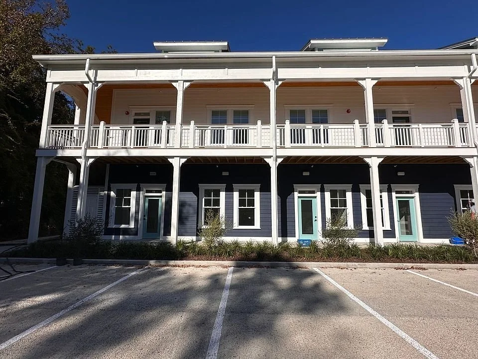 A two-story residential building with a large front porch, white railings, and dark blue siding. There are six doors on the ground floor and six windows on the upper floor, with a parking lot in front.