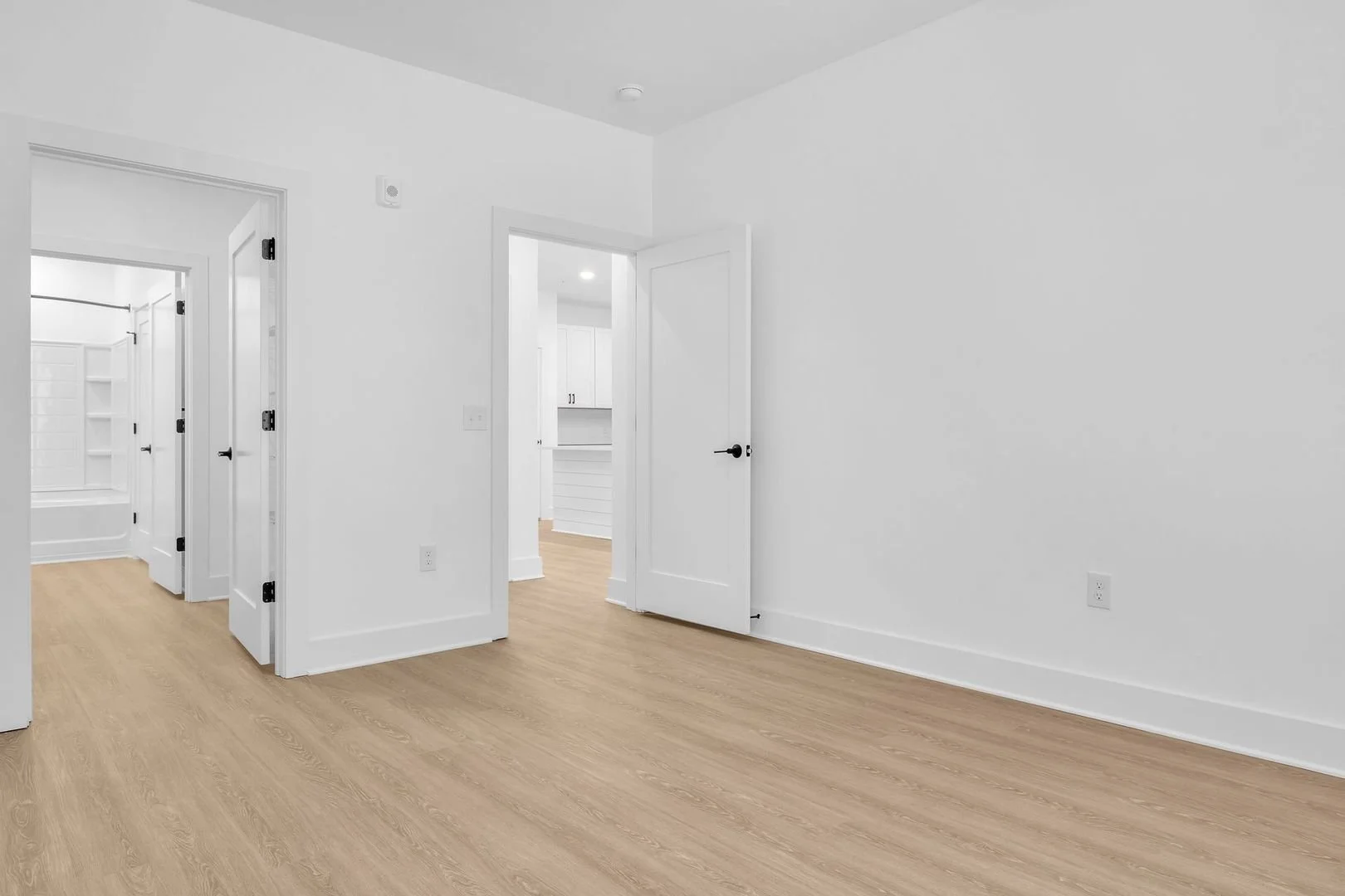 Empty room with white walls, wooden flooring, and open white doors leading to other rooms.