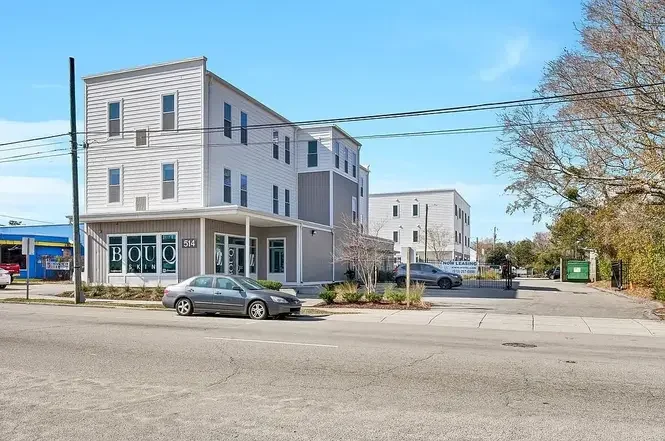 A three-story gray and white building on a city street, with a parking lot and a few cars in front, and trees along the sidewalk under a clear blue sky.
