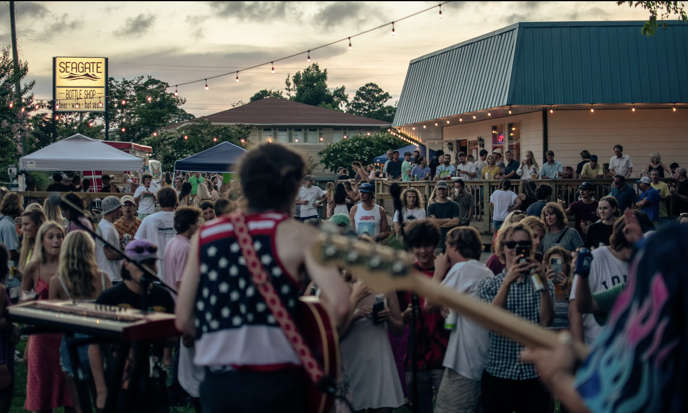 Outdoor music event with a crowd of people, a musician with an American flag-themed sleeveless shirt playing guitar in the foreground, and a Seagate bottle shop sign in the background during sunset.
