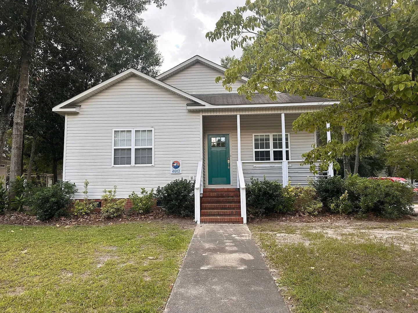 A beige multi-story house with a green door, a small front yard with bushes, and a driveway, surrounded by tall trees under a clear blue sky.