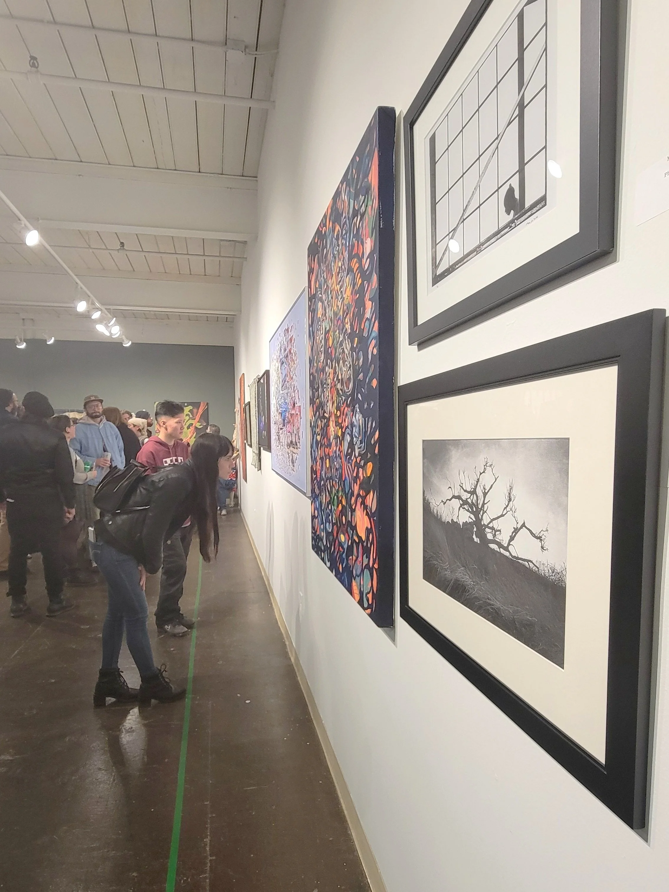 image of a gallery opening with paintings and photographs on the wall and people looking at the art