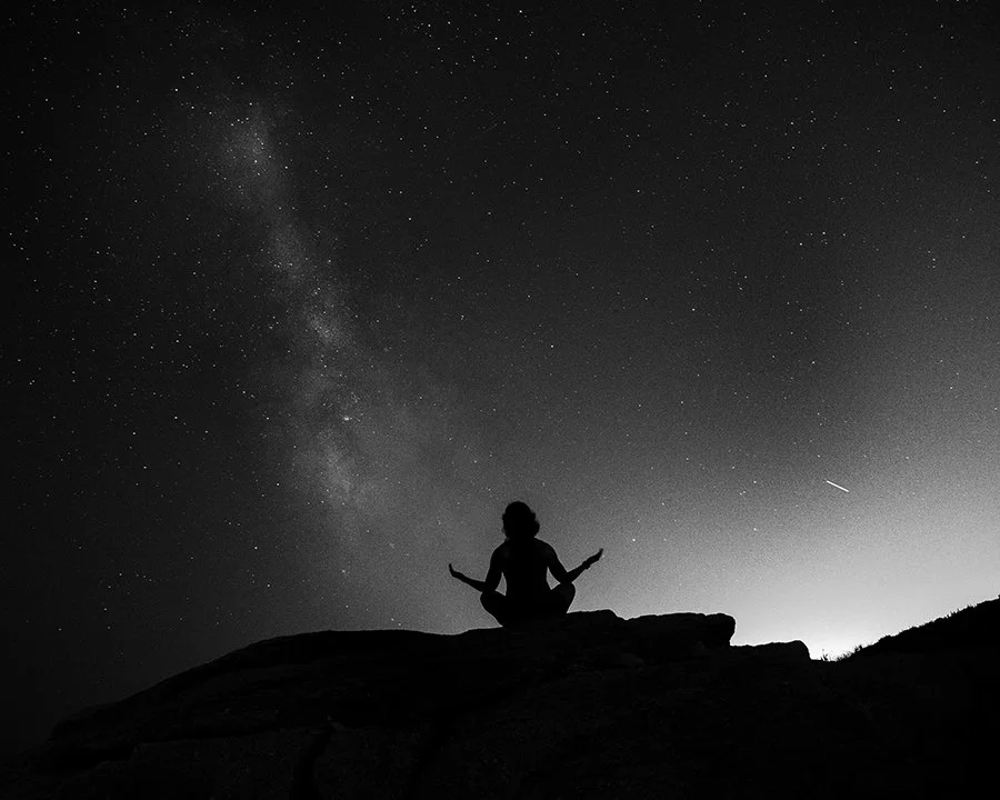 a silhouetted woman meditates under the night sky with the milky way and a shooting star in view