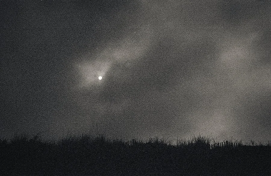 a grainy black and white photograph of the sun breaking through dark clouds over silhouetted sand dunes