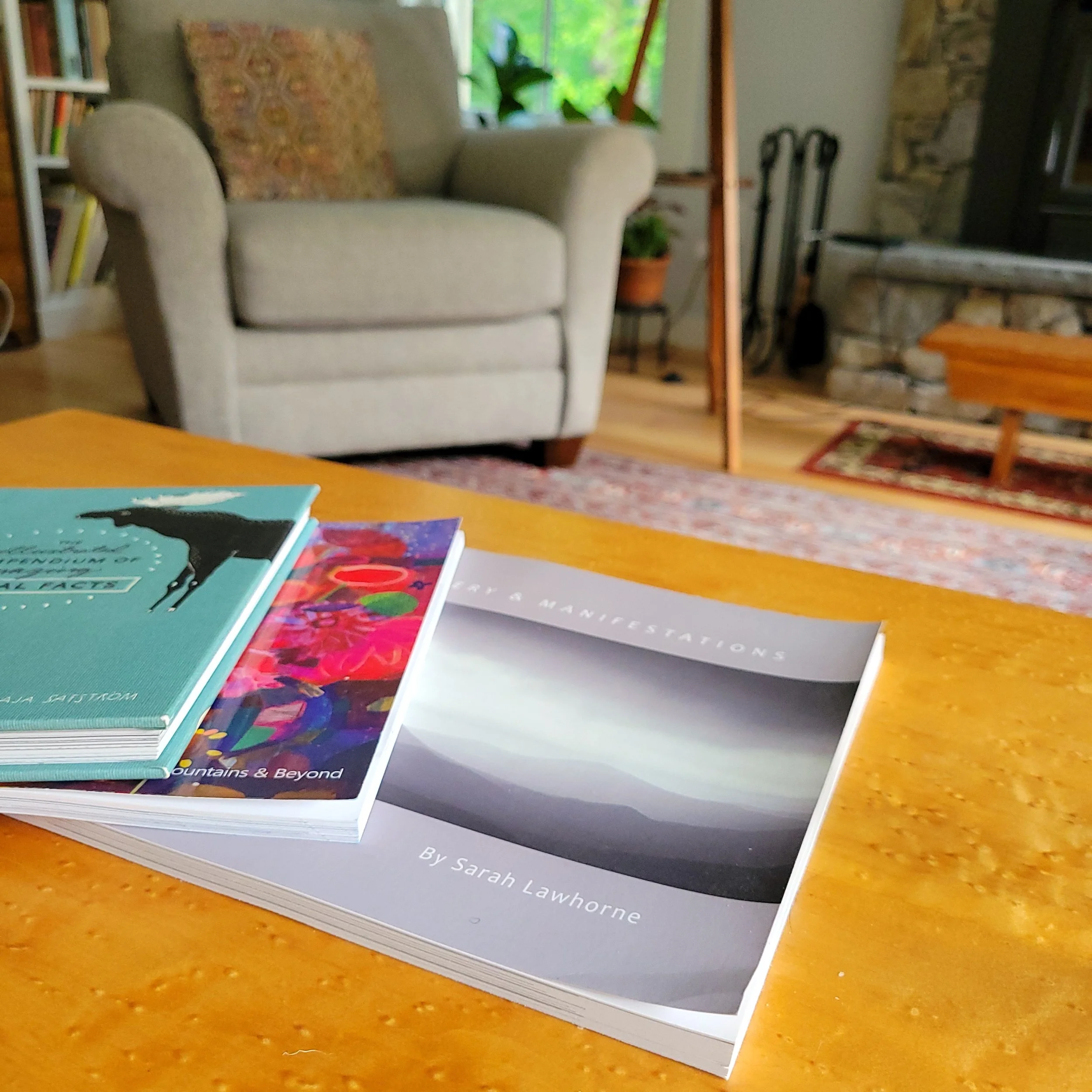a stack of books on a wooden table. in the background is a fireplace and comfortable chair in someone's living room