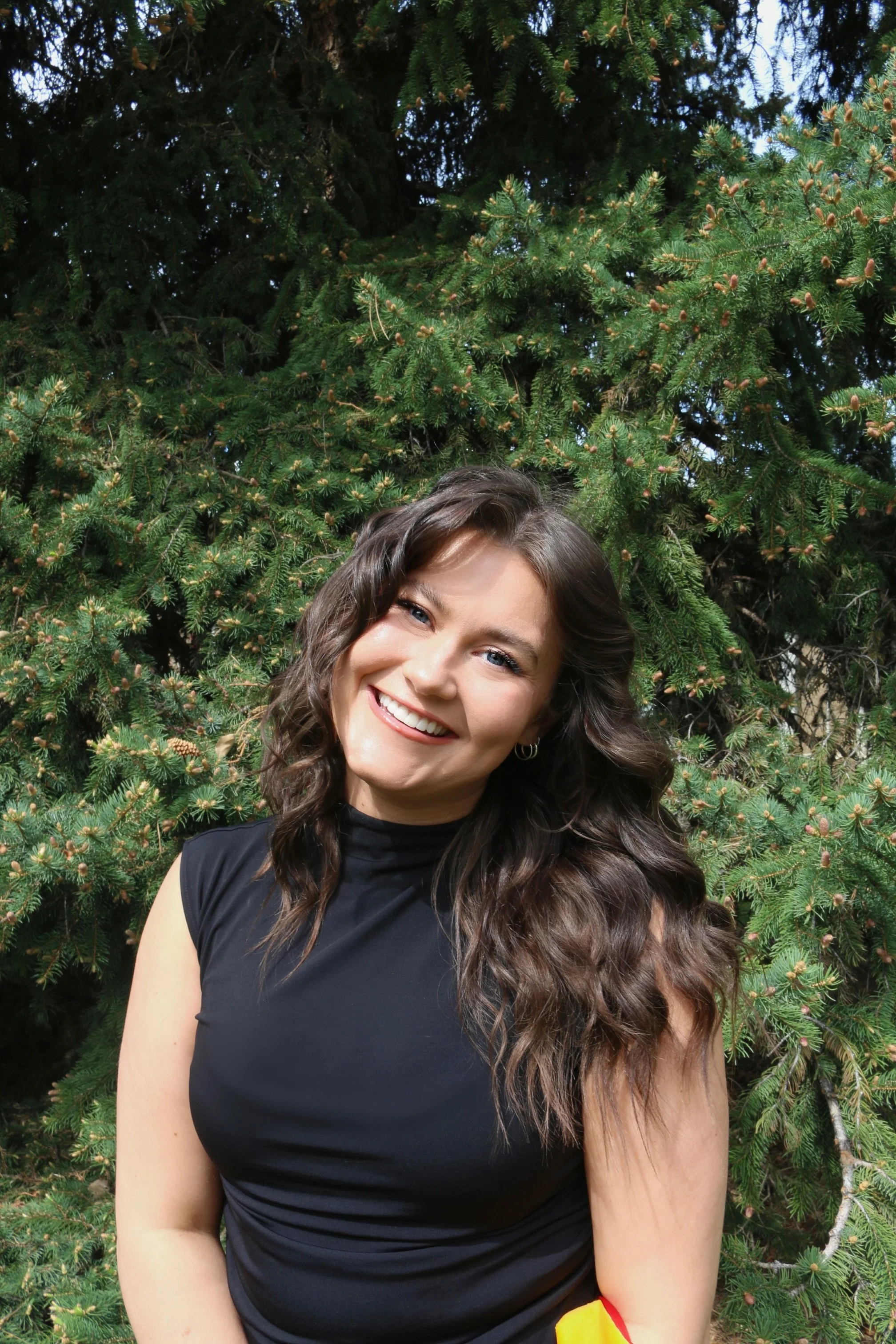 Smiling woman with long, curly brown hair and blue eyes standing outdoors in front of a lush green pine tree.