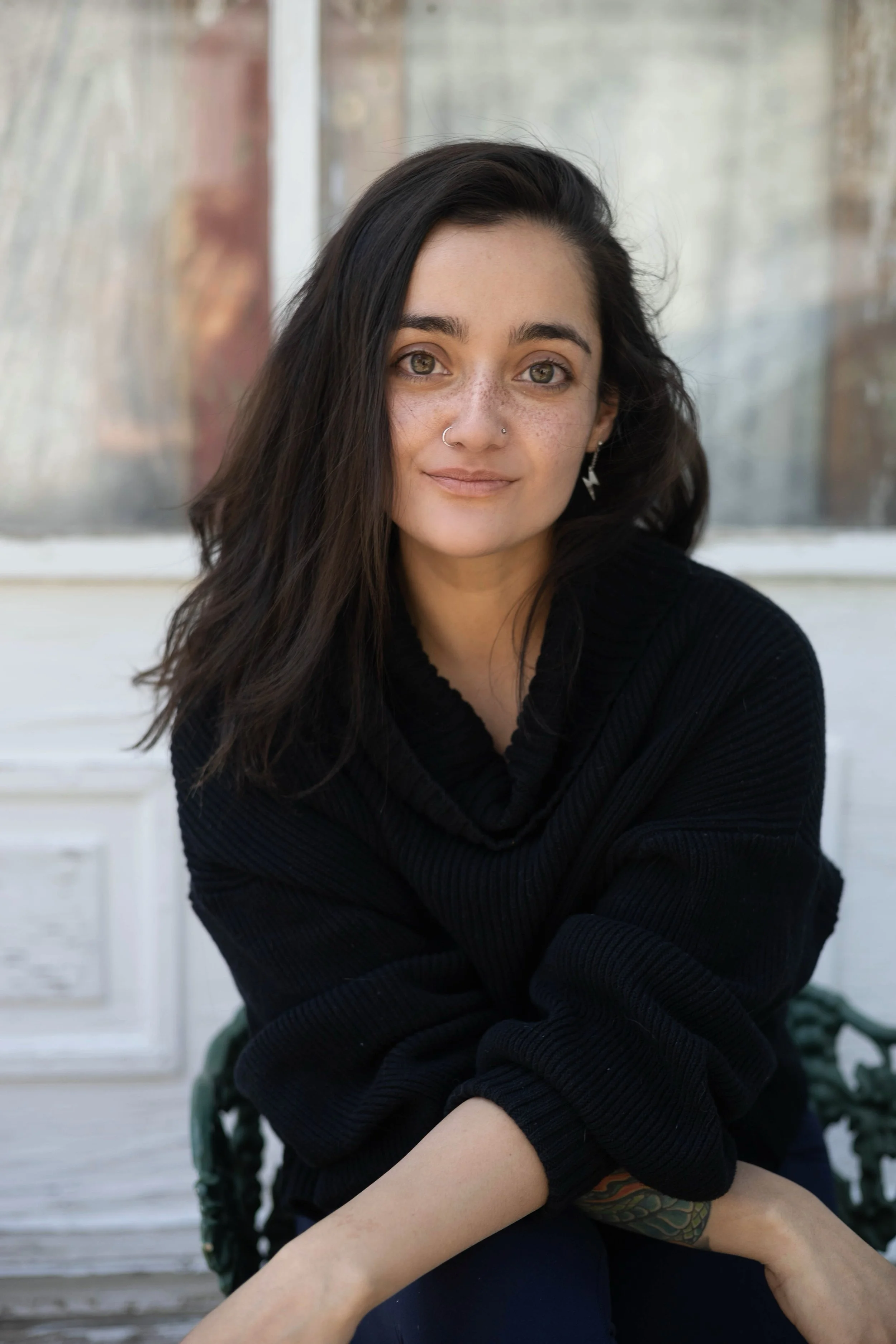 A woman with long dark hair, light complexion, and freckles, wearing a nose ring, earrings, and a black top, sitting outdoors with a blurred background of a window and brick wall.
