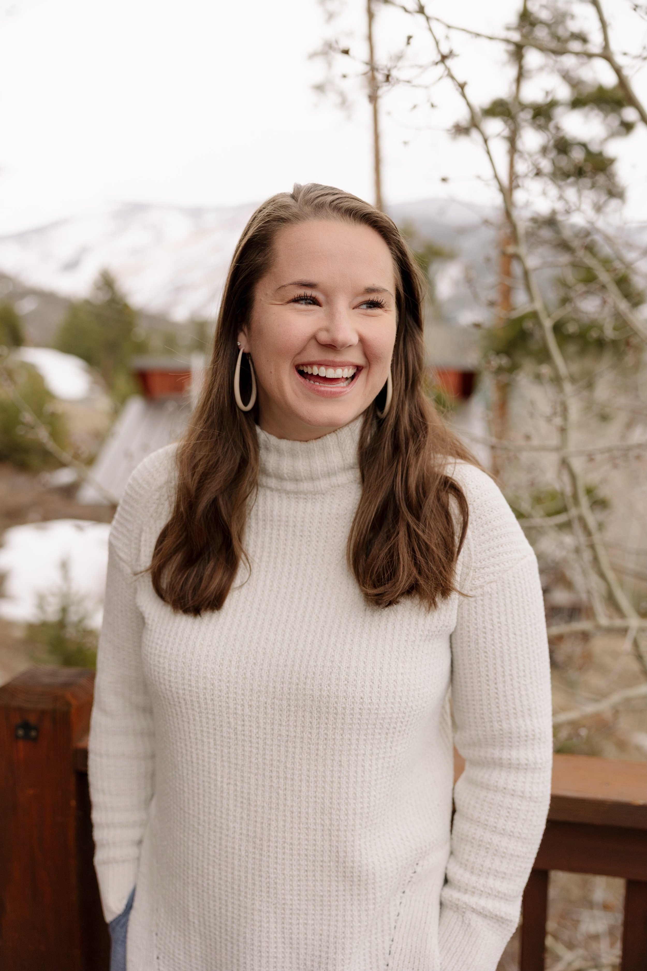 Woman with long brown hair wearing a white turtleneck sweater and hoop earrings, outdoors in a mountain setting with snow and trees in the background.