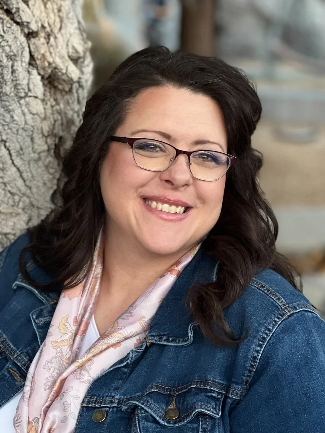 A woman with dark wavy hair and glasses smiling at the camera, leaning against a tree in an outdoor setting.