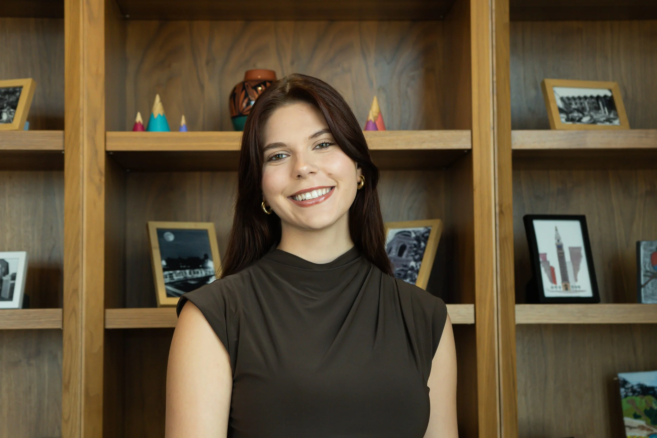 A woman with long brown hair and gold hoop earrings smiling in front of a wooden bookshelf filled with framed photos and decorative items.
