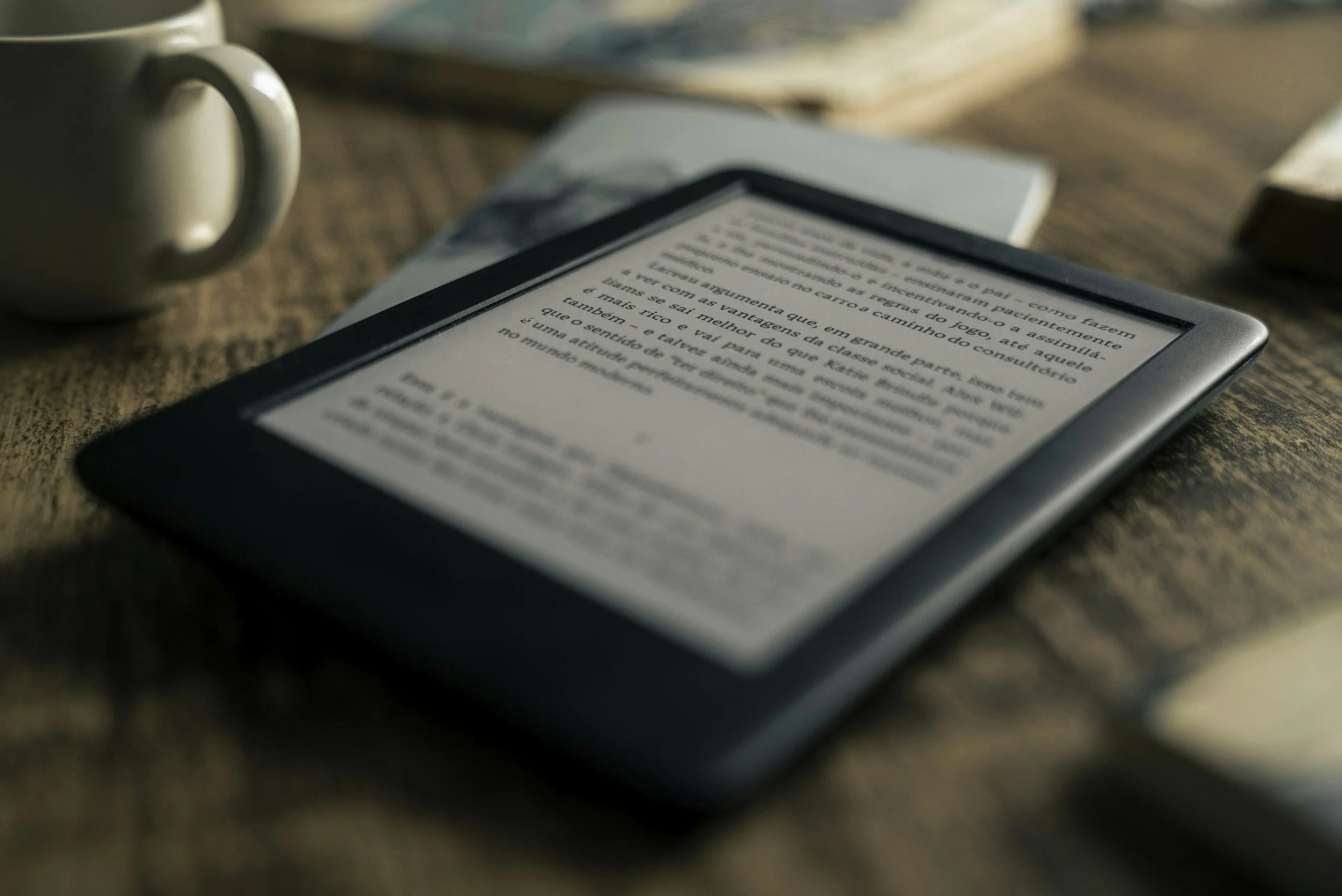 Close-up view of an e-reader displaying text in Portuguese, placed on a wooden surface near a ceramic mug and some books.