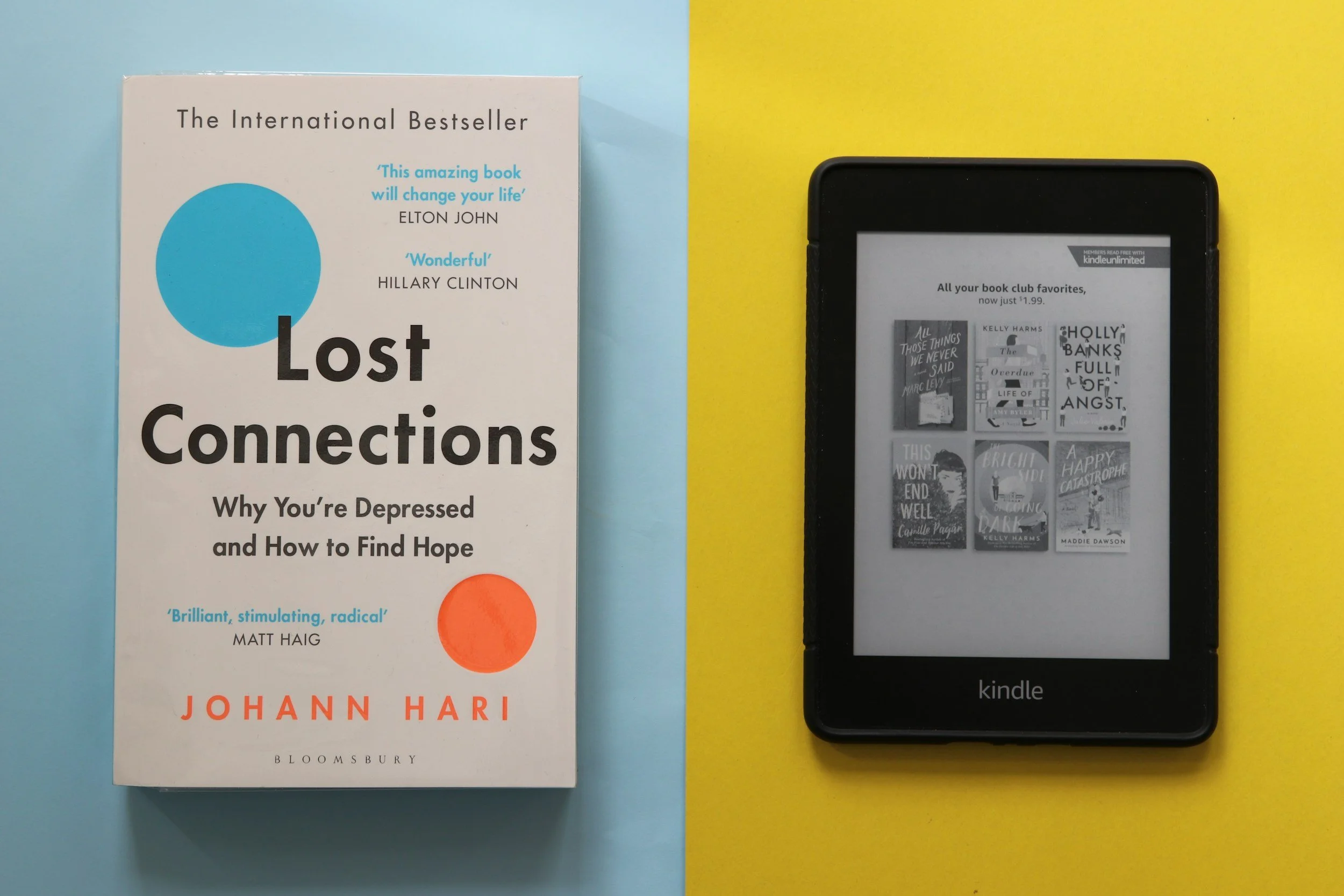 A physical copy of the book "Lost Connections" by Johann Hari laid on a light blue background, with a Kindle e-reader displaying pages from various books on a yellow background, partially visible on the right side of the image.