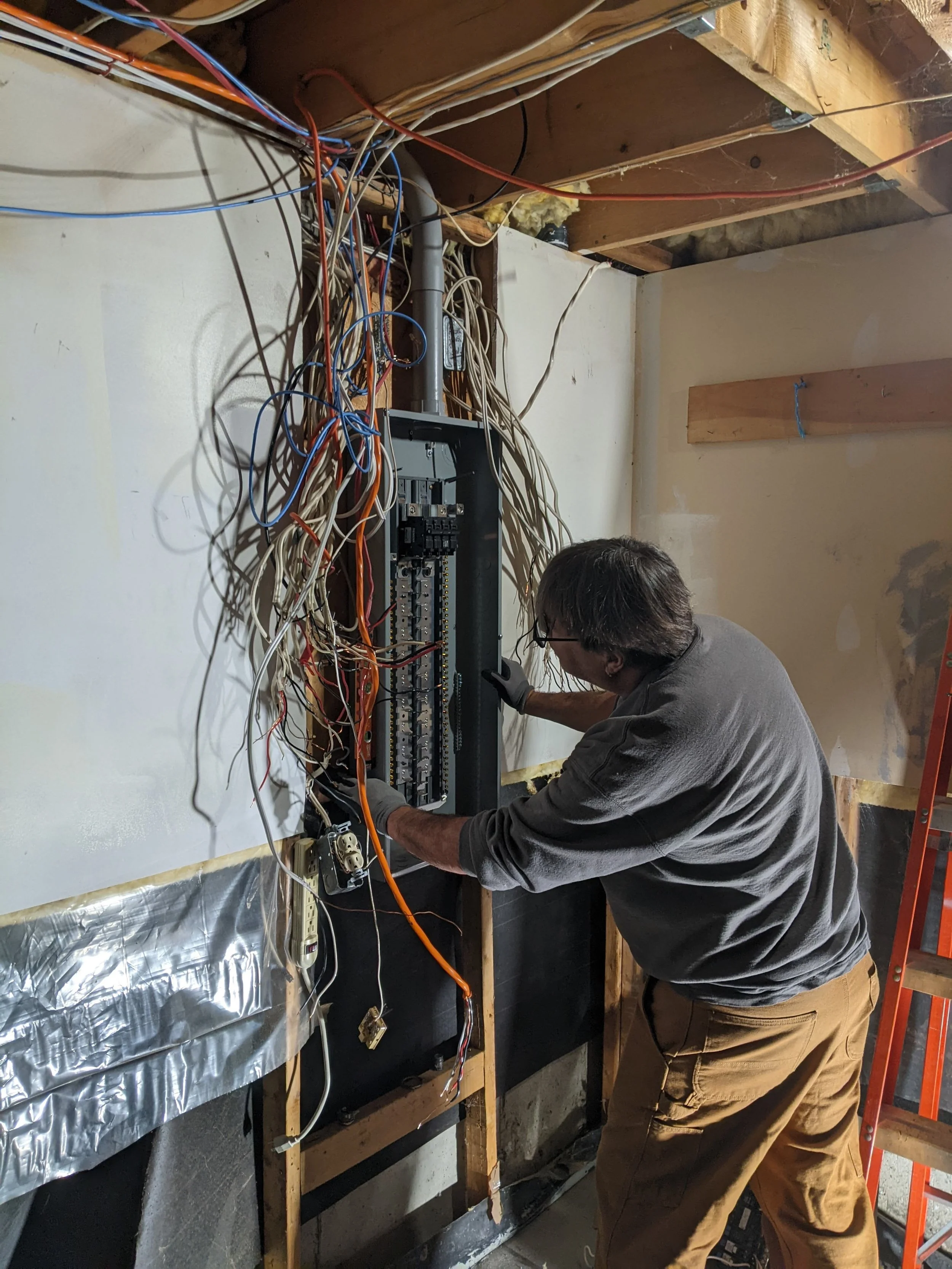 A man working on an electrical panel with various wires connected. The panel is mounted on a wall in a partially finished room with exposed wooden beams and insulation.