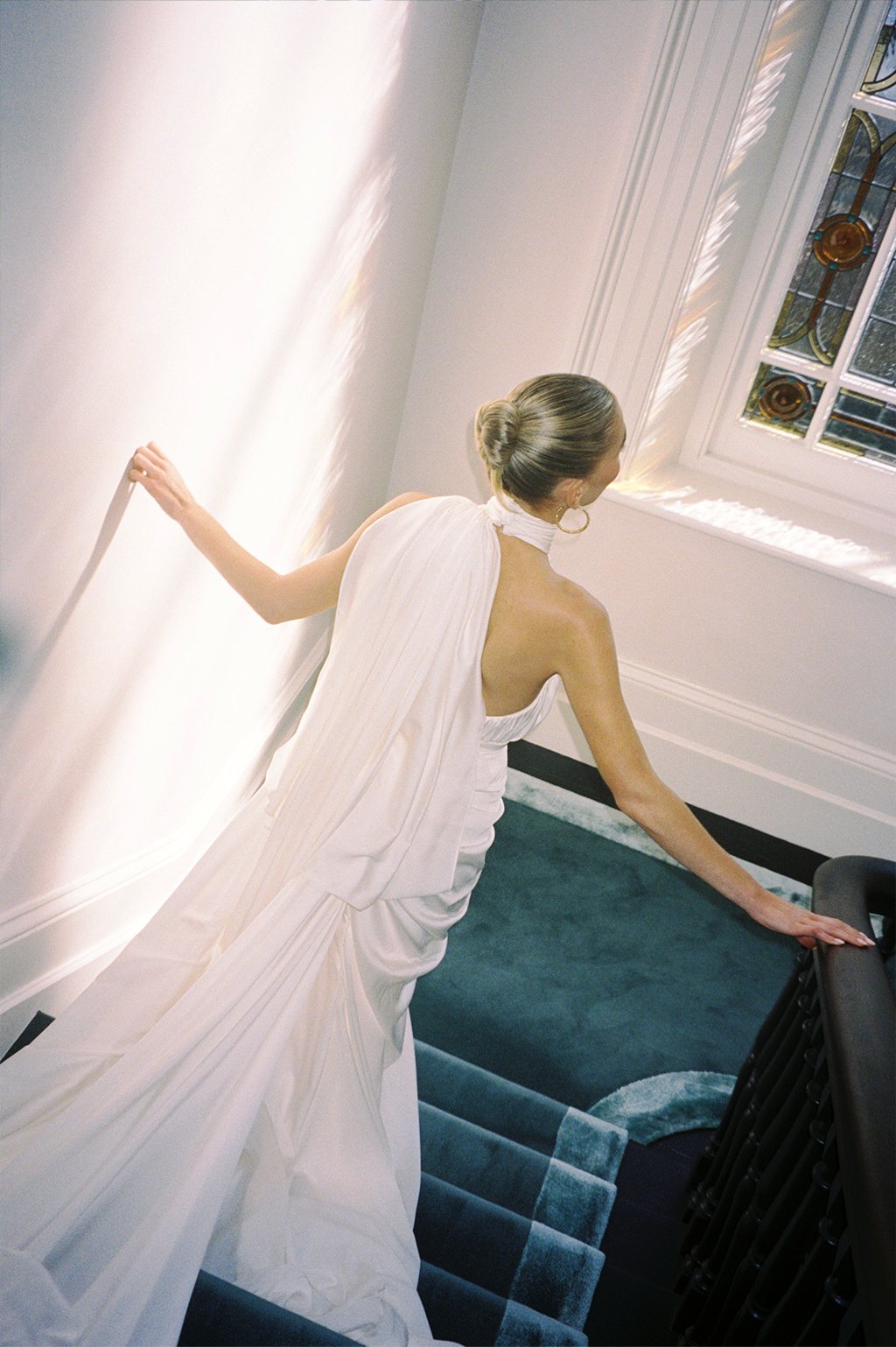 A woman in a white dress, with her hair in a bun, standing on a staircase near a window, with sunlight casting reflections on the wall.
