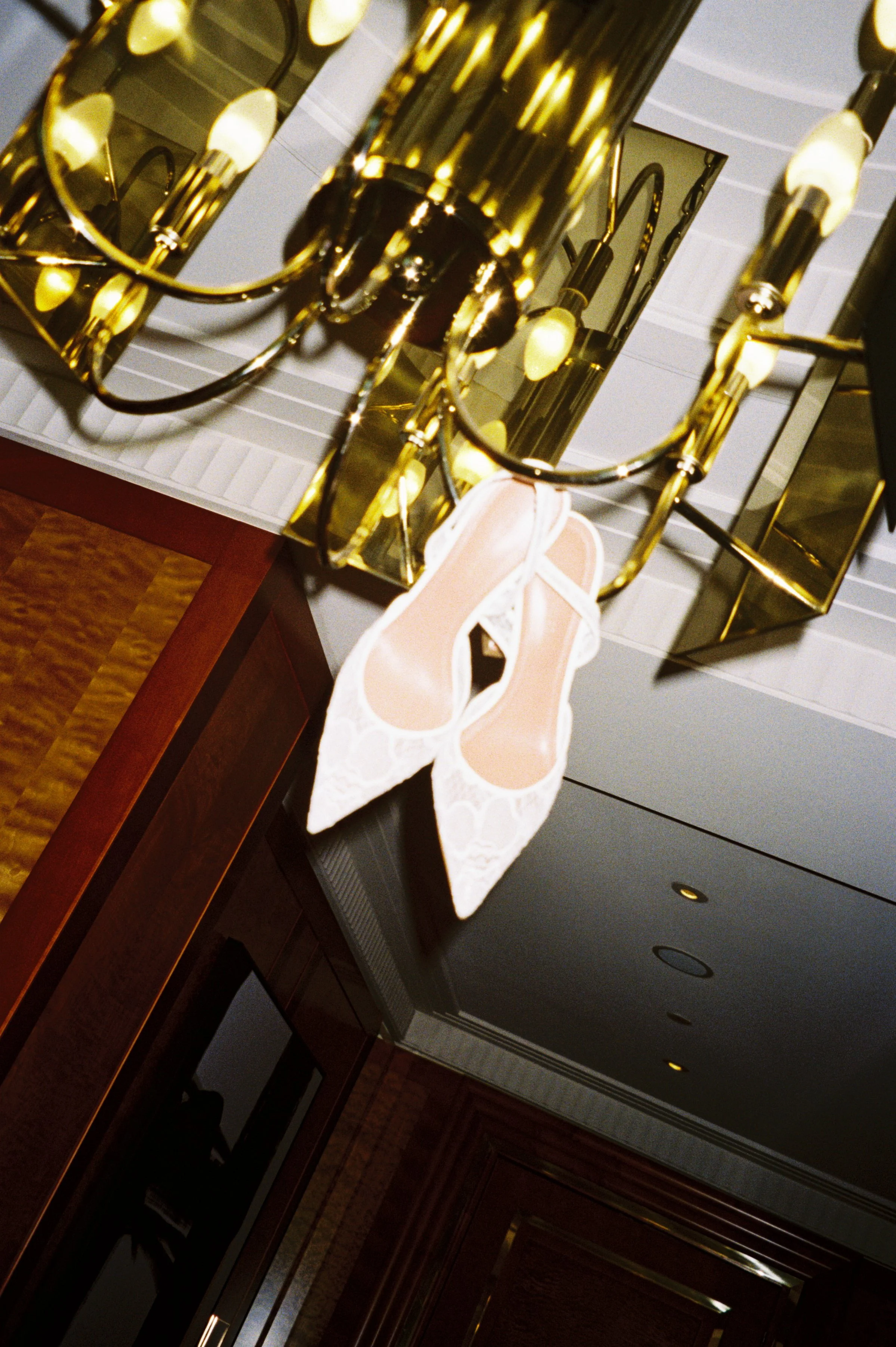 A pair of white lace high-heeled shoes hanging under a golden chandelier in a formal room.
