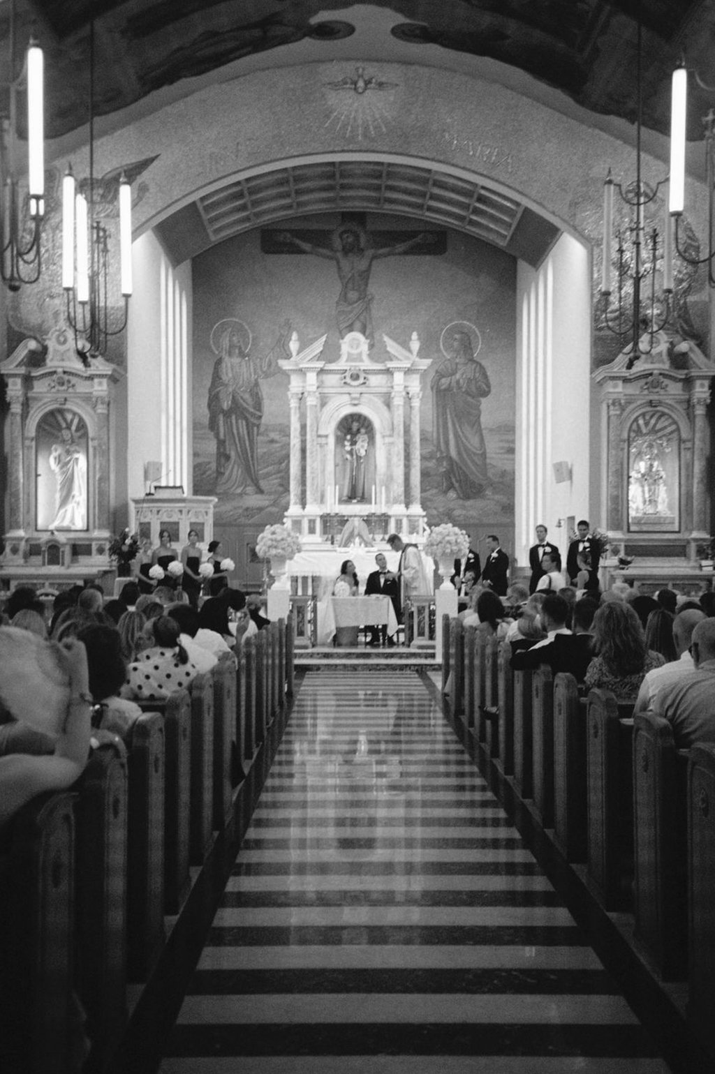A church wedding ceremony with the bride and groom kneeling at the altar, surrounded by attendants and guests seated in pews, with a large religious painting of Jesus crucifixion on the background.