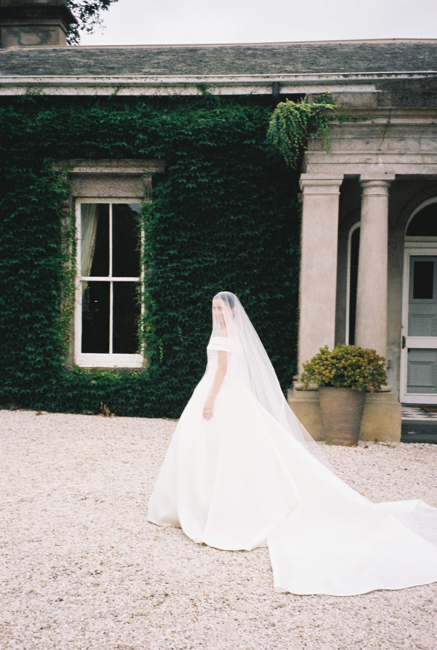 A bride in a white wedding dress and veil standing outdoors in front of an ivy-covered building with a window and columns.