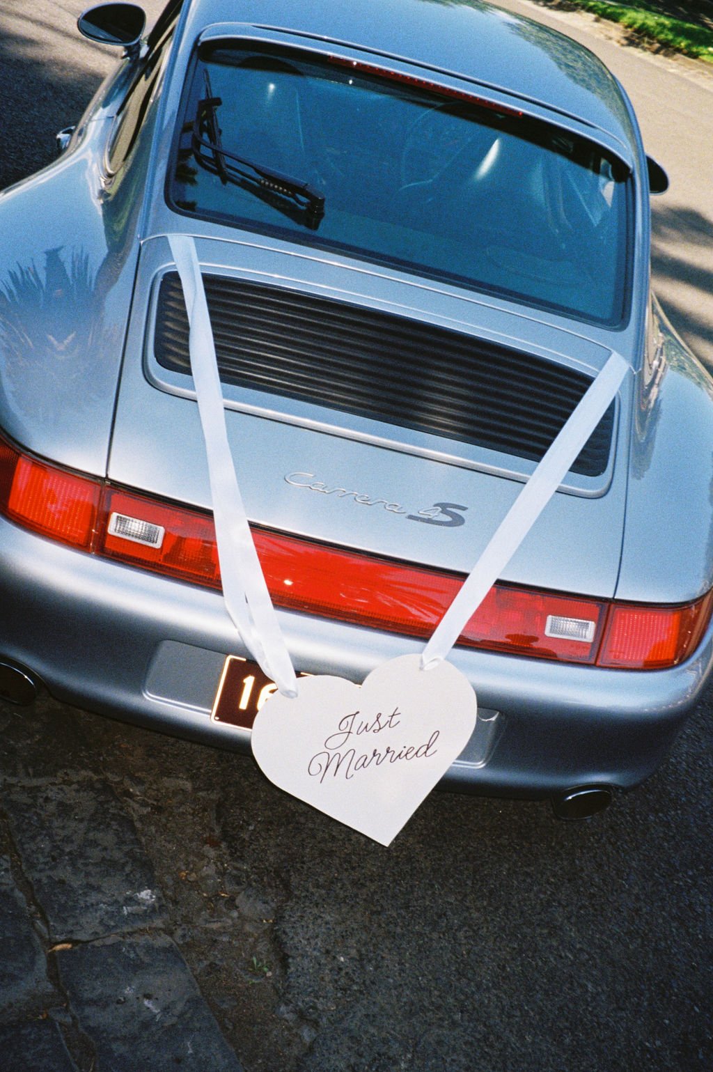 A silver Porsche Carrera 5 parked outdoors with ribbons and a heart-shaped sign that reads 'Just Married' attached to the back.