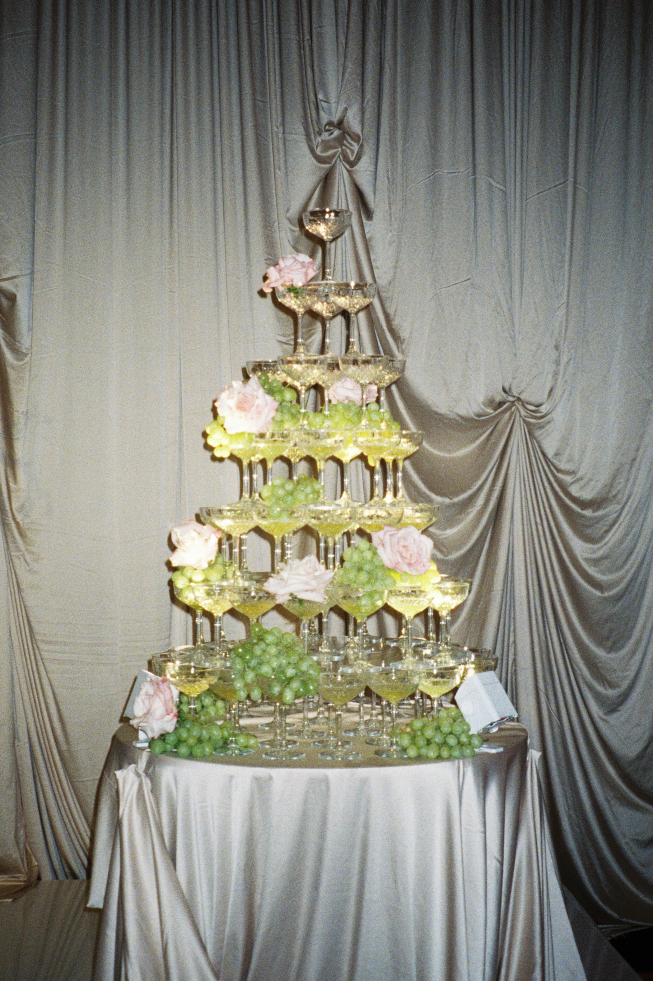 A pyramid of champagne glasses decorated with pink roses and green grapes, set against a gathered satin backdrop.