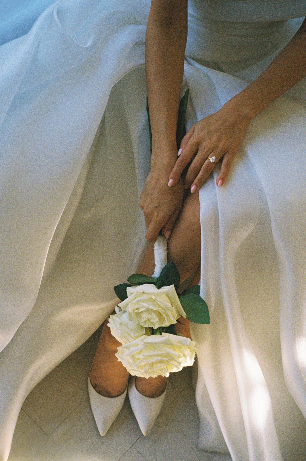 Person wearing white heels, a white dress, and a ring on their finger, holding a bouquet of white roses, sitting on a white silk fabric.