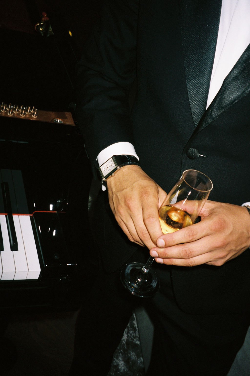Close-up of a man in a tuxedo holding a glass of champagne, with a piano and a foosball table in the background.