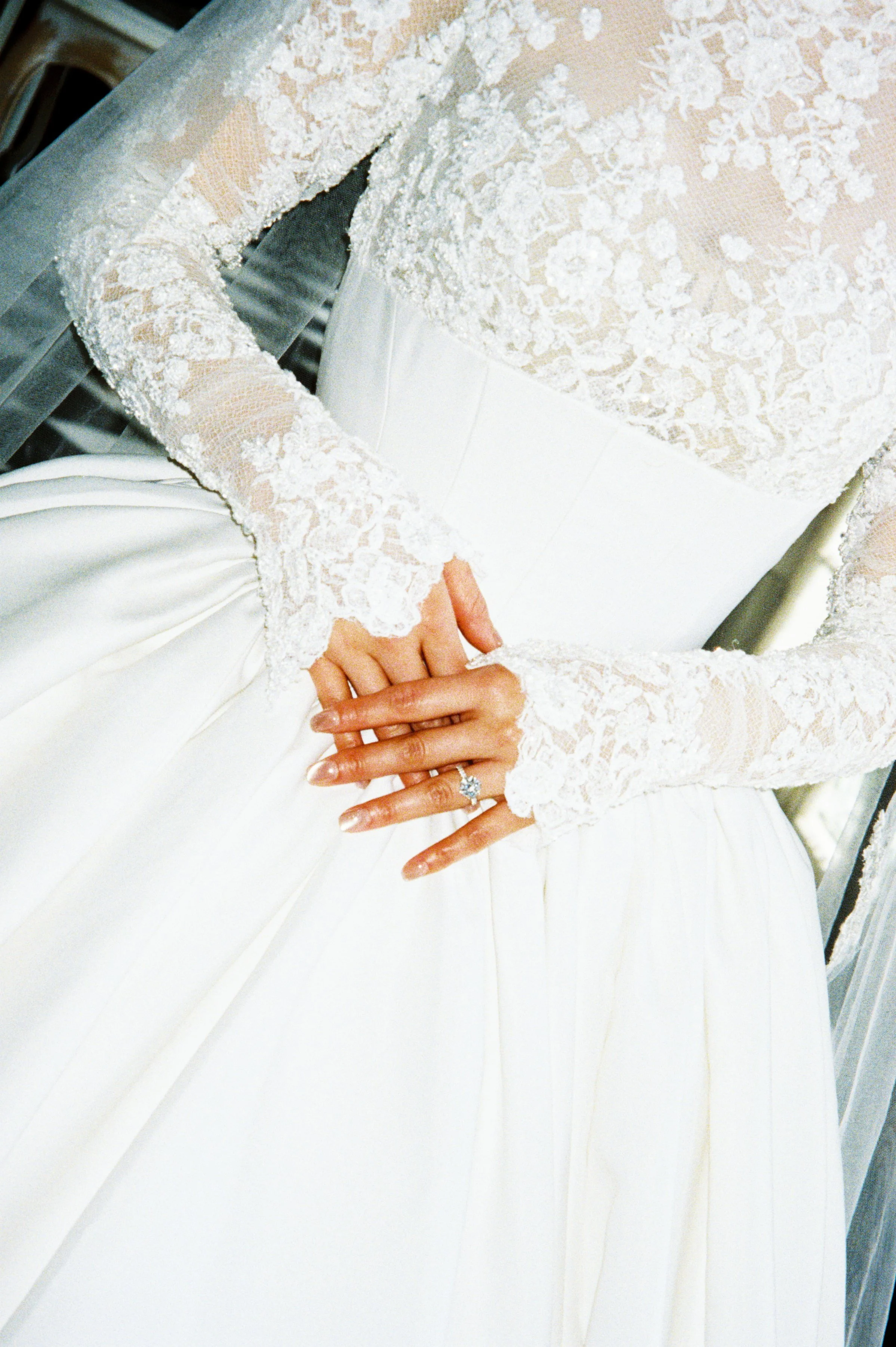 Close-up of a bride in a white wedding dress with lace sleeves, showing her hand with a diamond engagement ring and wedding band resting on her waist.