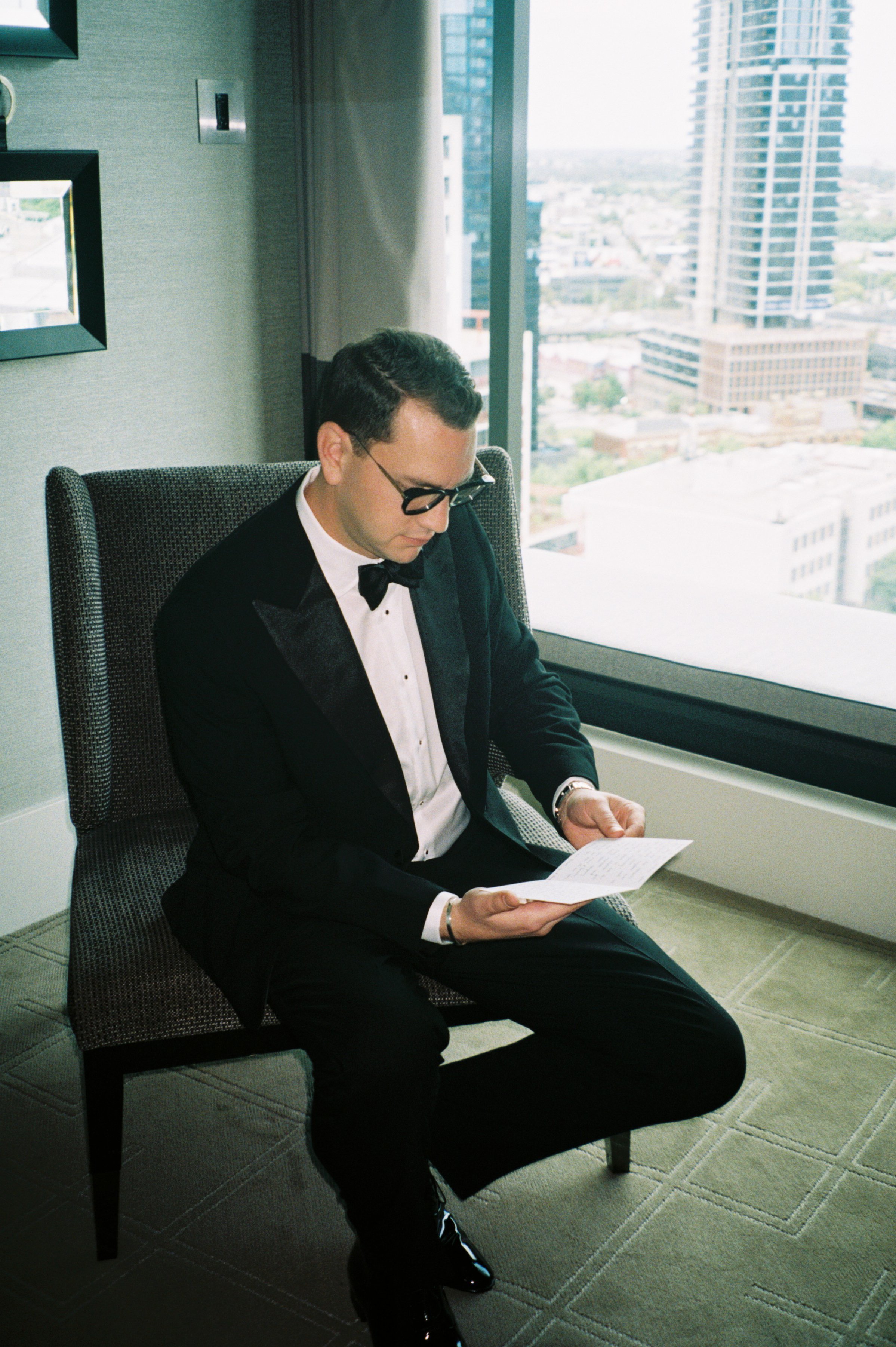 A man wearing a tuxedo, sitting in a chair by a large window, reading a letter with a city skyline view behind him.