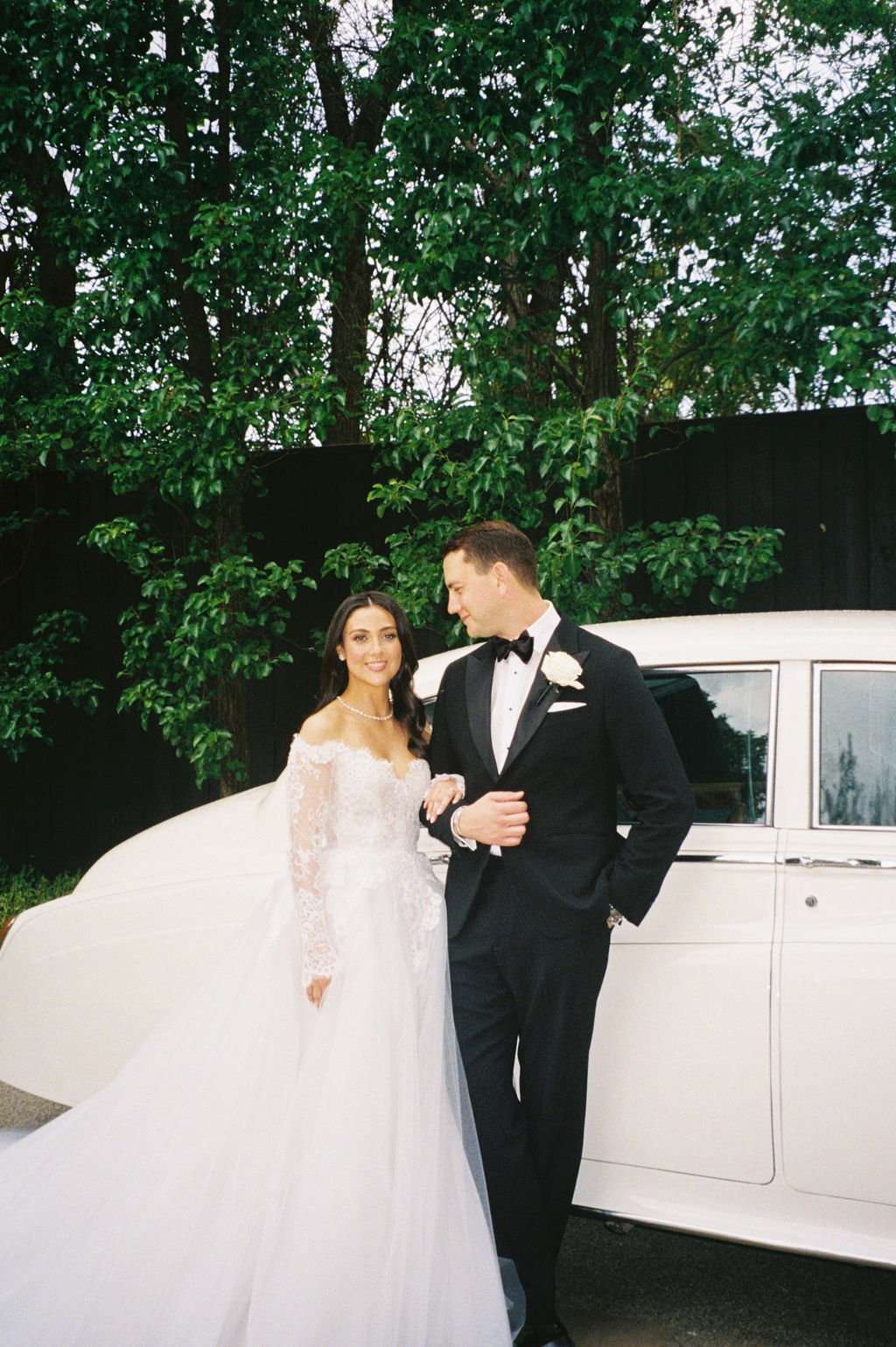 A bride and groom standing in front of a vintage white car outdoors, with green trees and a black fence in the background. The bride is wearing a white wedding gown with lace details, and the groom is dressed in a black tuxedo with a bow tie and a wh