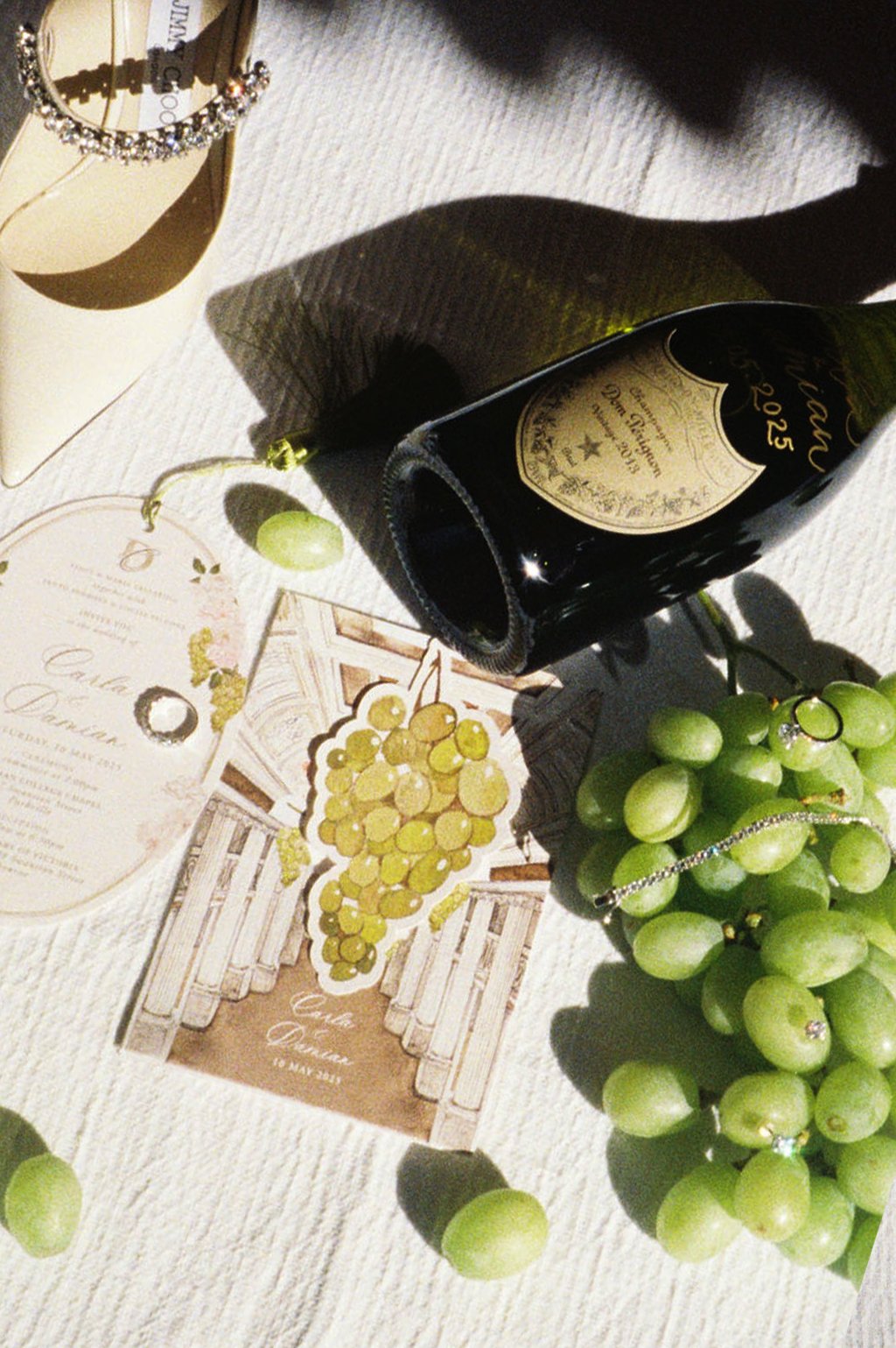 A bottle of champagne, green grapes, wedding rings, wedding invitation, and a greeting card on a white textured surface.