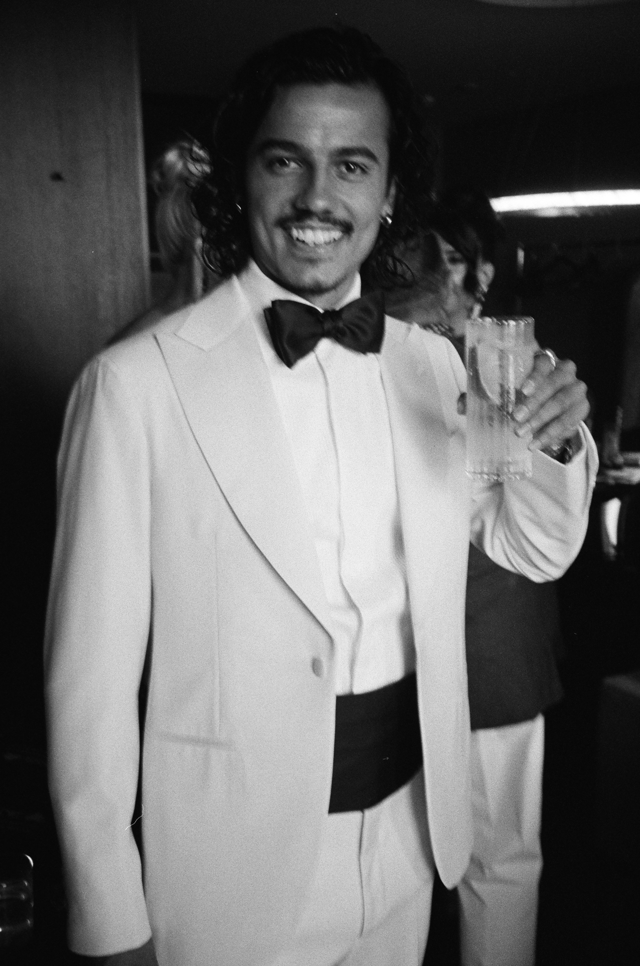 A man dressed in a white tuxedo and black bow tie, smiling, holding a glass, at a formal event in black and white photography.