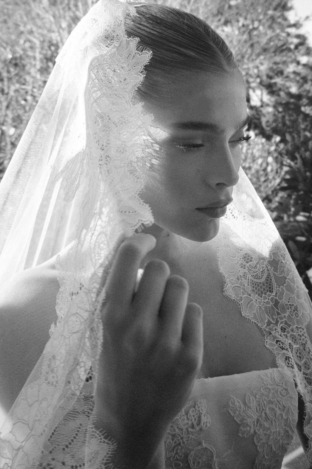 A black and white photo of a woman wearing a lace wedding veil, with her hand gently touching the veil near her face.