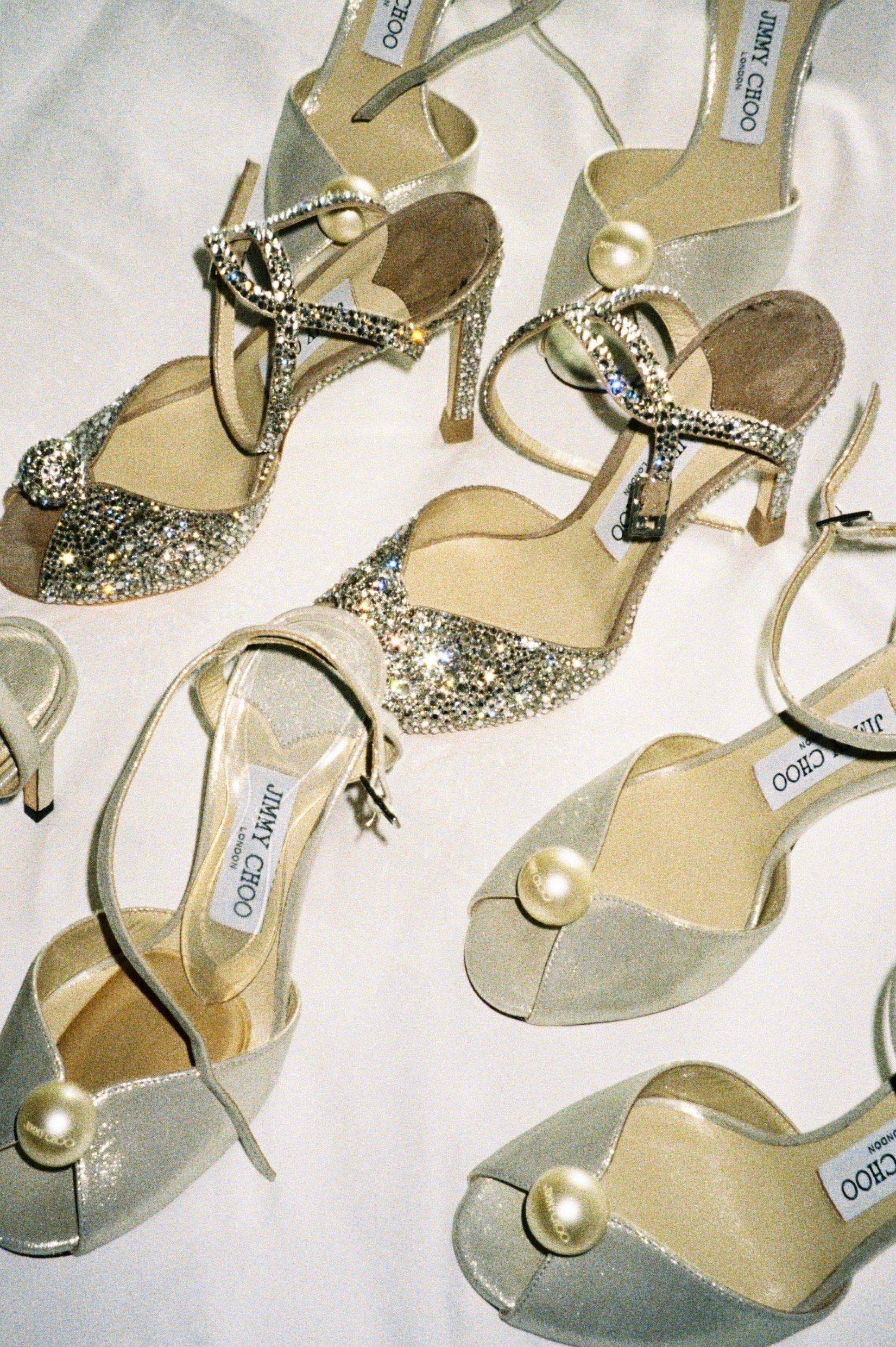 A display of glamorous high-heeled shoes, predominantly in light colors with glitter and pearl accents, some with ankle straps and decorative bows, branded 'Jimmy Choo'.