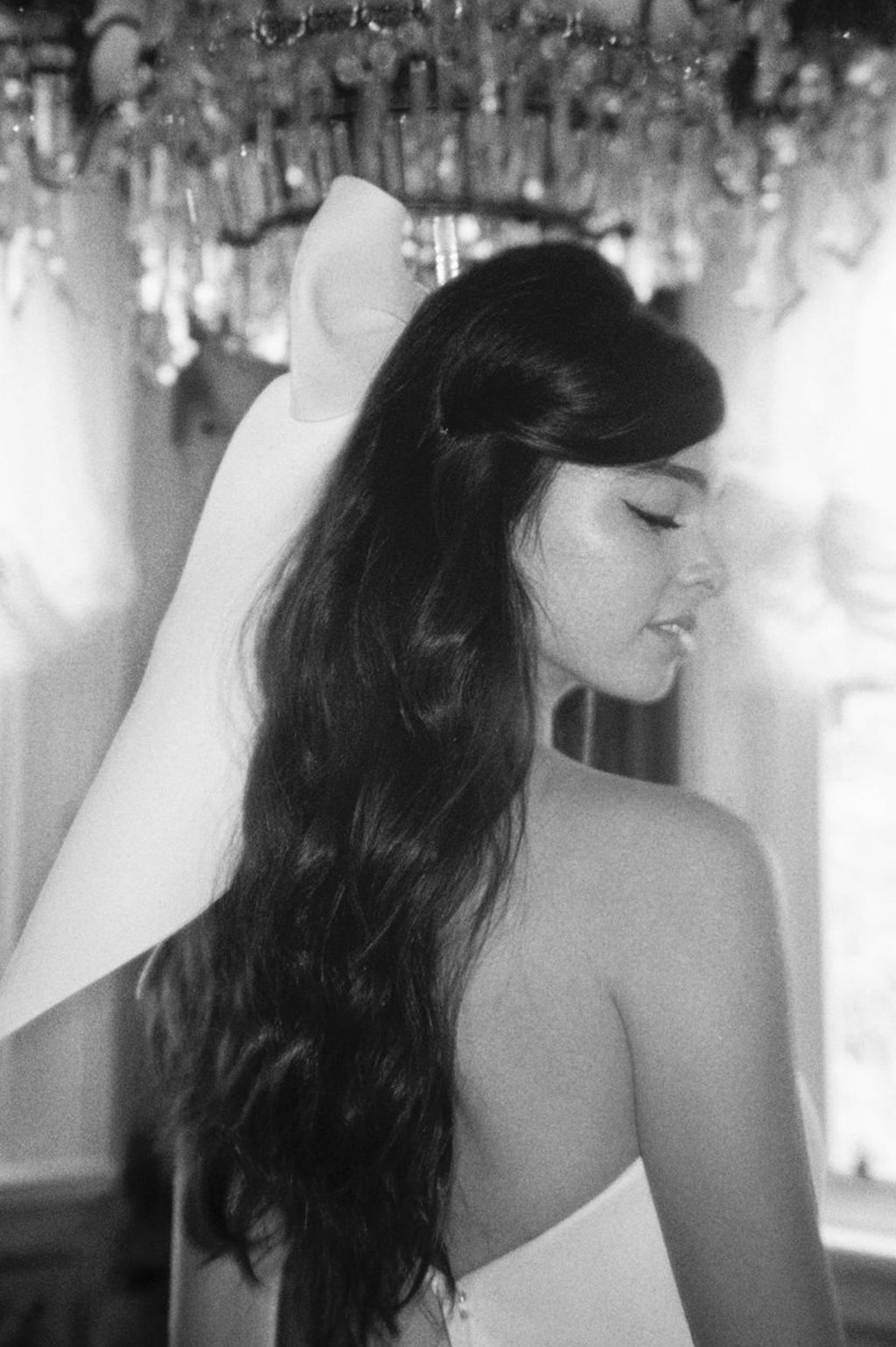 Black and white photo of a woman with long, wavy dark hair, closed eyes, and her head slightly tilted downward. She is wearing a strapless top, and there is a large feathered hat or accessory on her back, with a blurred background showing a decorated