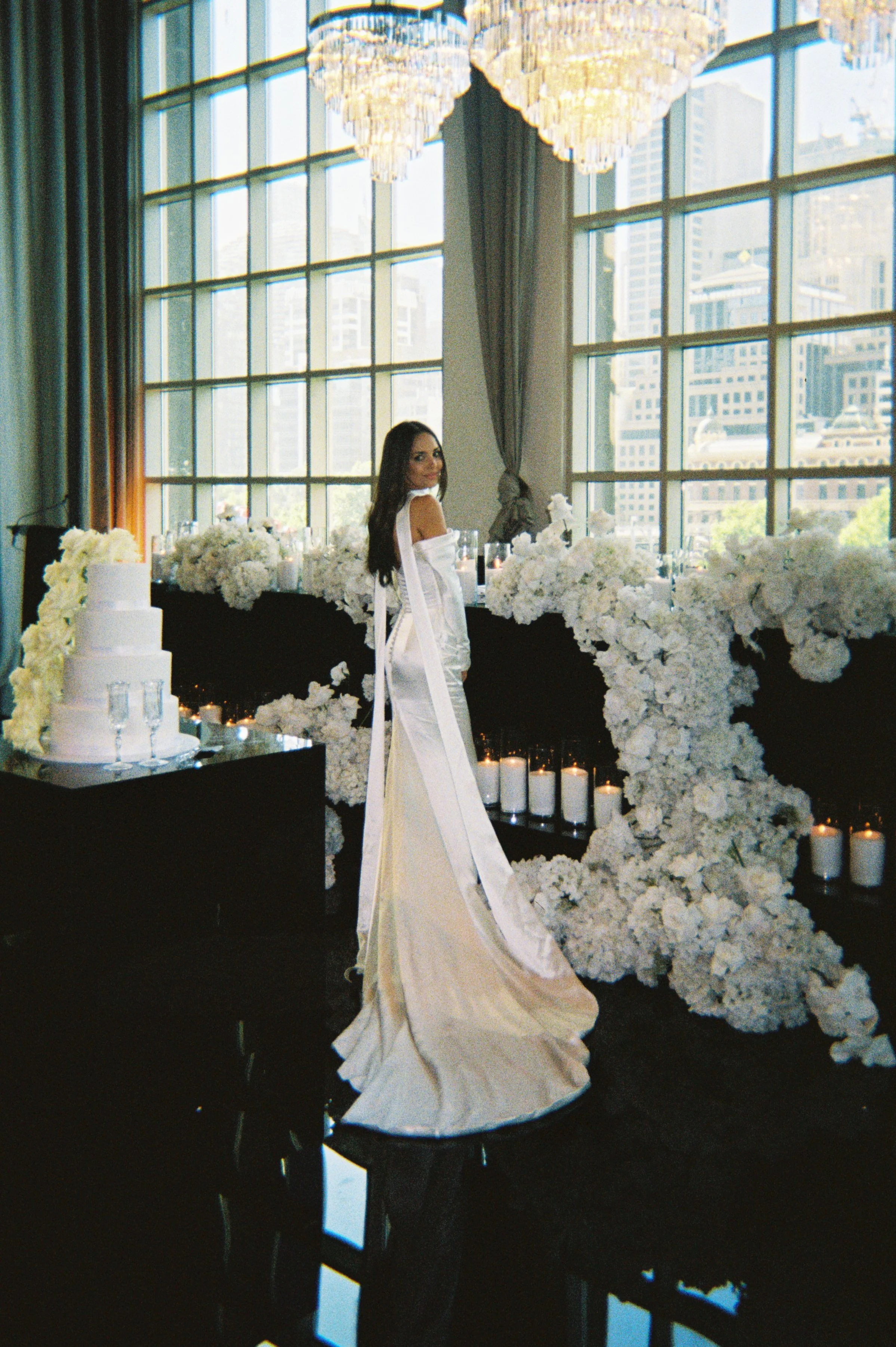 A woman in a white gown with long train at a wedding reception, standing in front of a floral and candle display near a large window in a high-rise building.