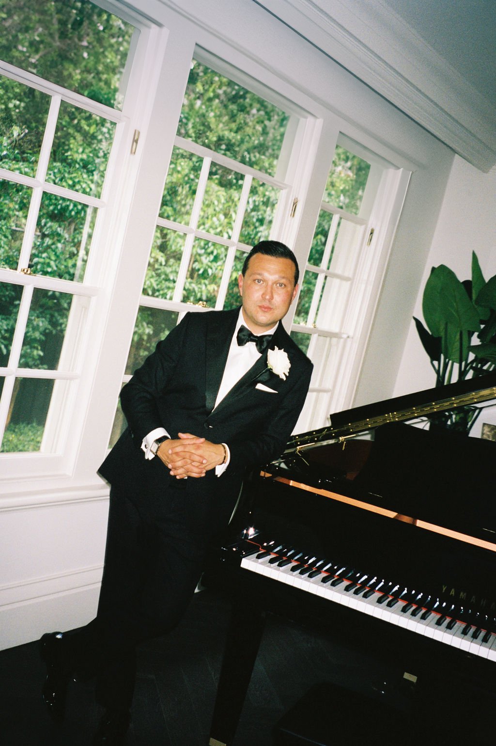 A man in a tuxedo with a white boutonniere standing beside a grand piano in a well-lit room with large windows and green trees outside.