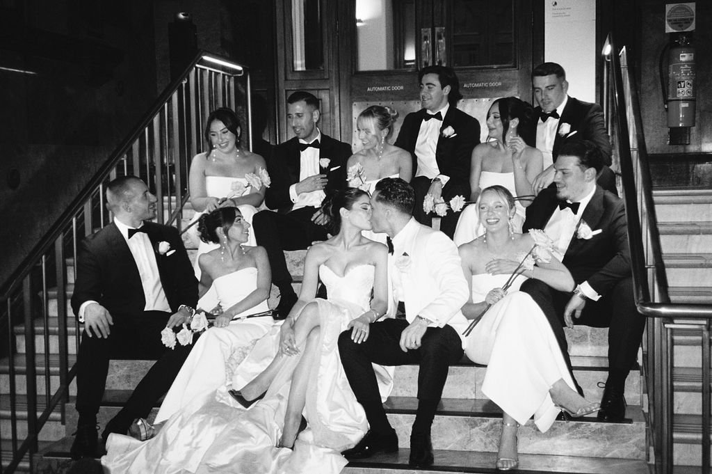 Black and white photo of a wedding party sitting on staircase, with the bride and groom kissing in the center.
