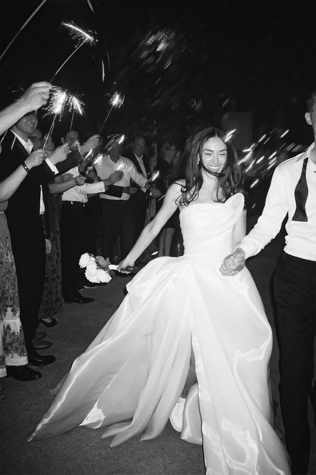 A bride in a strapless wedding gown holding hands with a groom in a tuxedo, smiling as they walk through a crowd at night, with guests holding sparklers.
