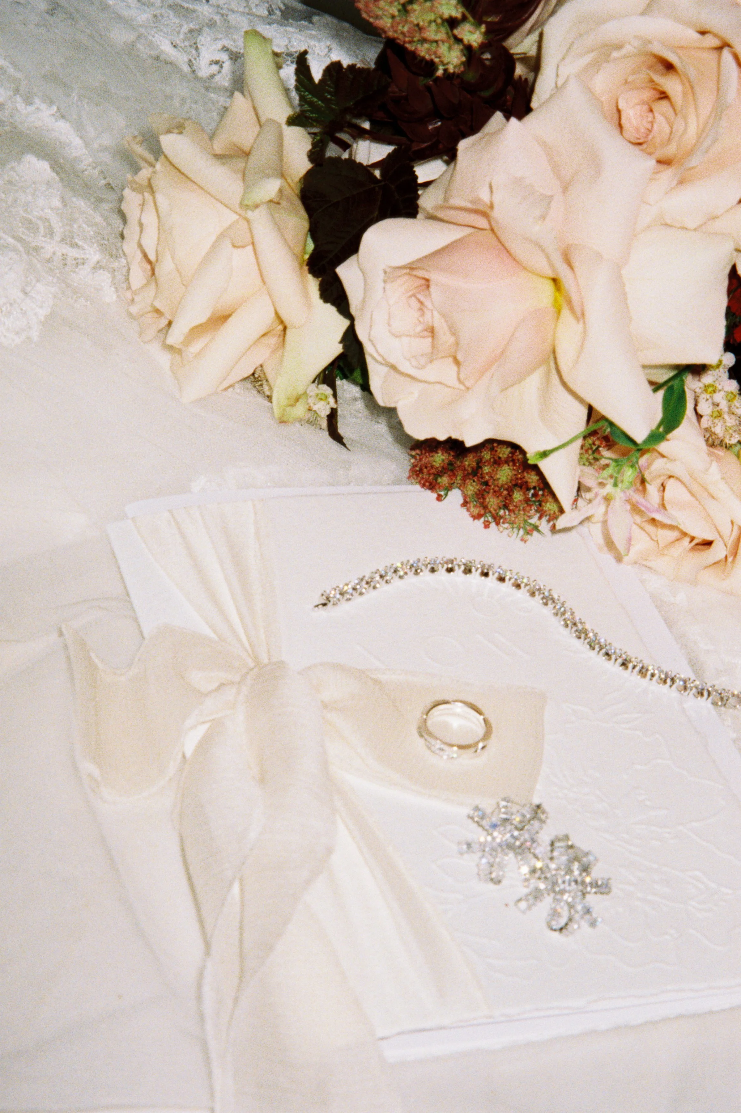 Wedding ring, jewelry, and a flower arrangement on a white tablecloth, with pale pink roses and greenery in the background.
