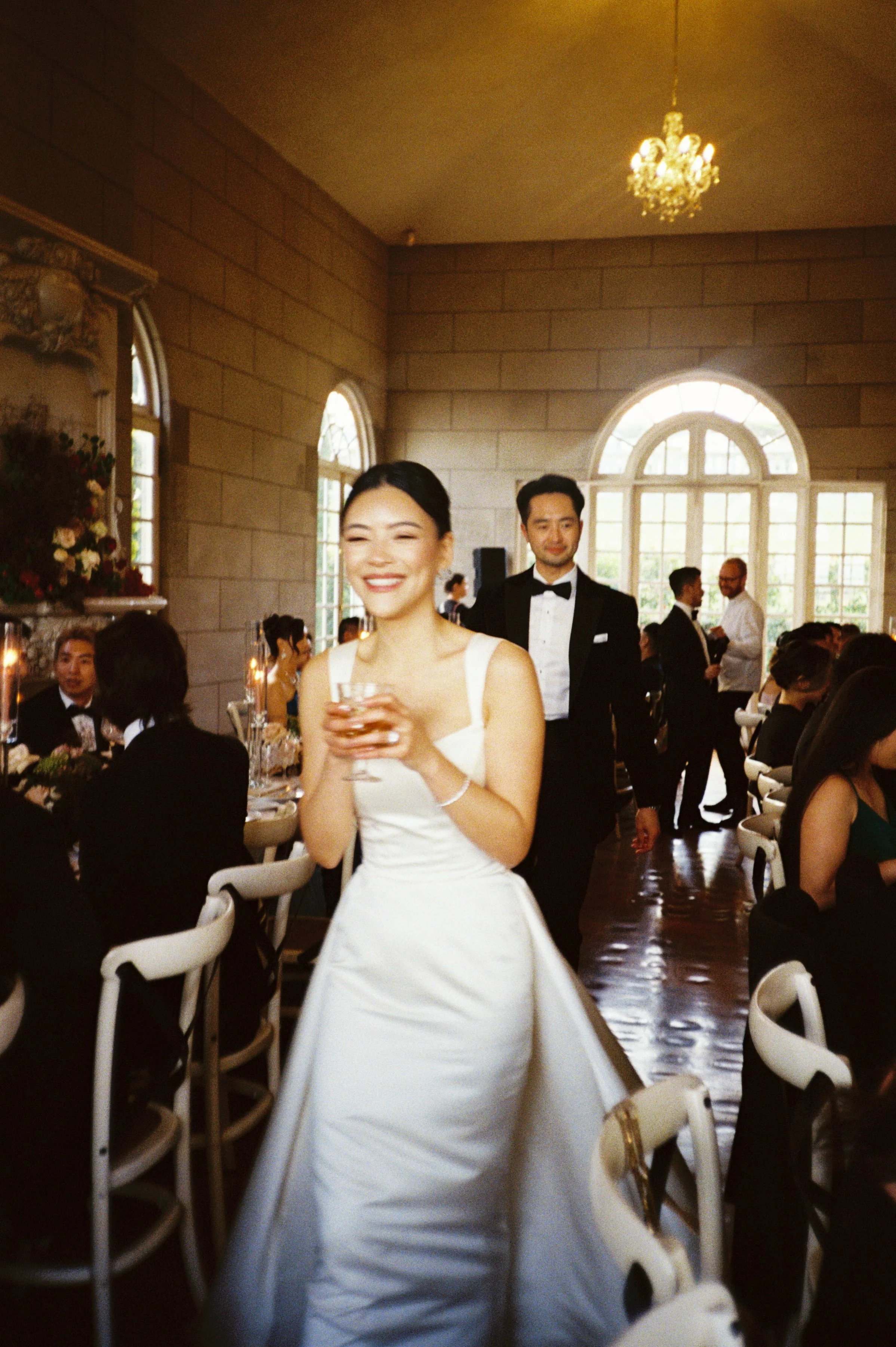 A bride in a white wedding dress smiling and holding a glass, walking through a decorated reception hall filled with seated guests and elegantly dressed people, with large windows and a chandelier overhead.