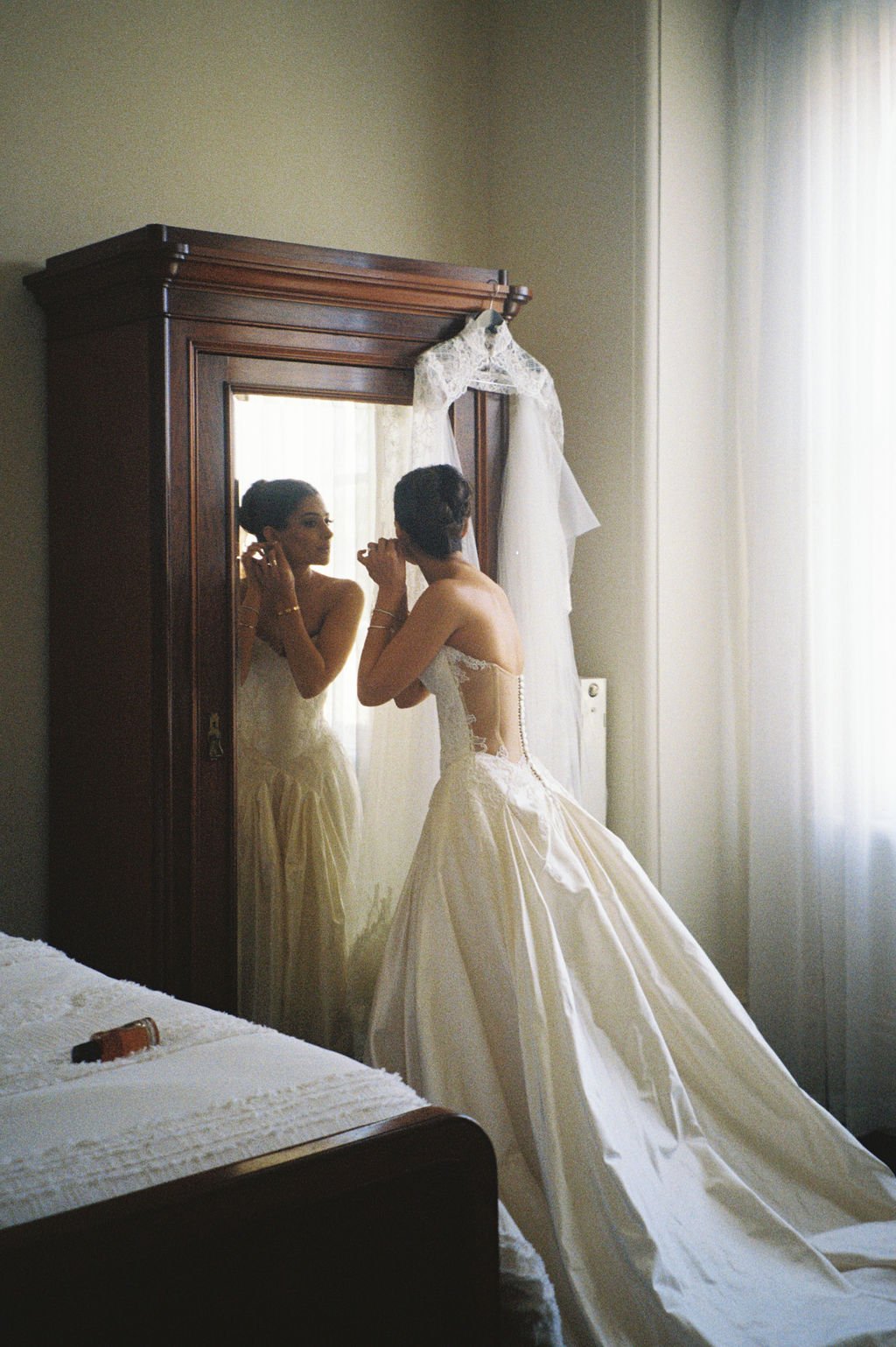 A bride in a wedding dress adjusting earrings in front of a mirror, with her reflection visible. The wedding dress has a fitted bodice with lace details and a full skirt. A white veil hangs on a wooden wardrobe, which is partially visible in the back