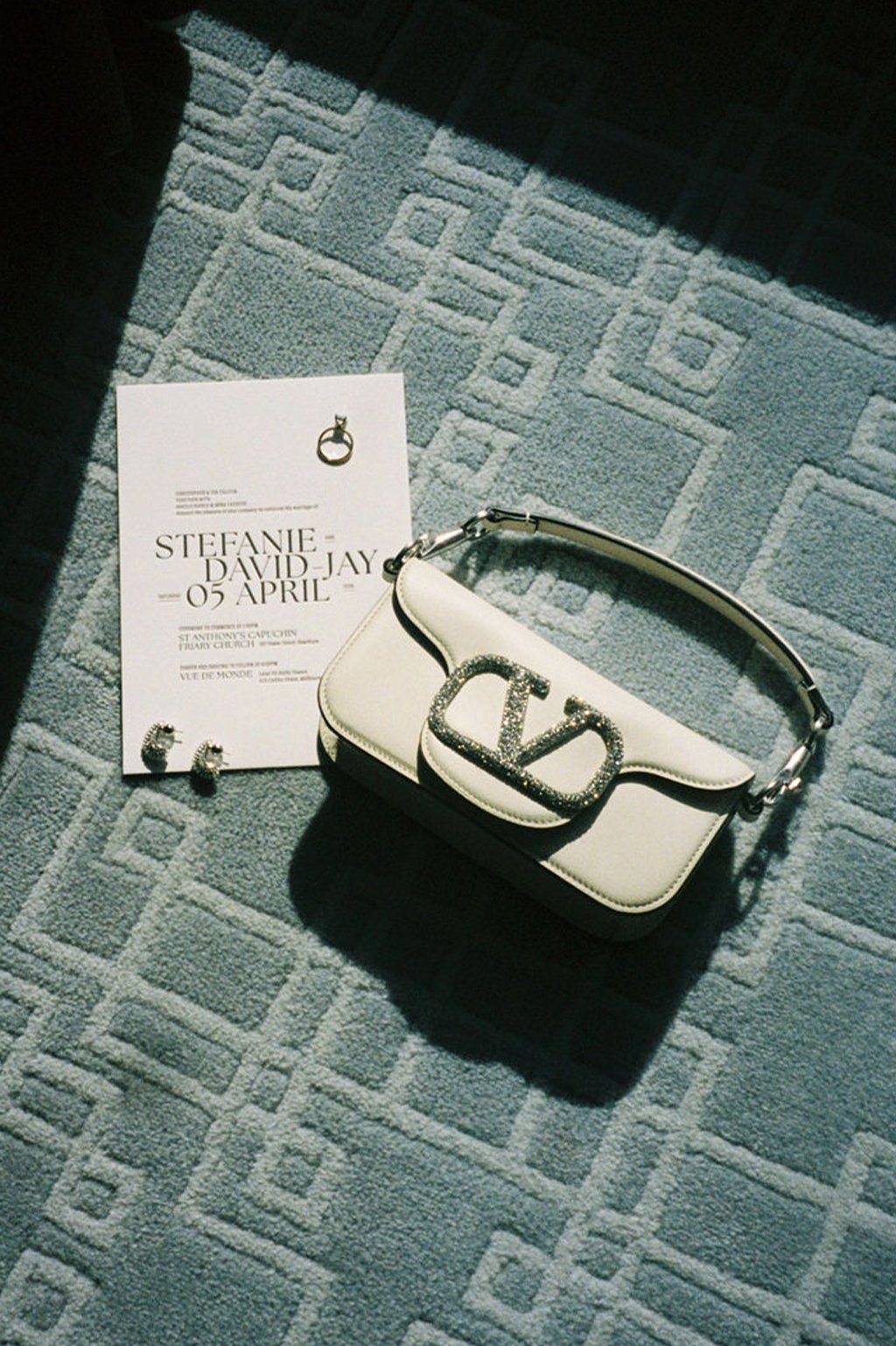 A white Valentino bag with a large V logo on the front, a pair of earrings, and a wedding invitation on a patterned carpet.